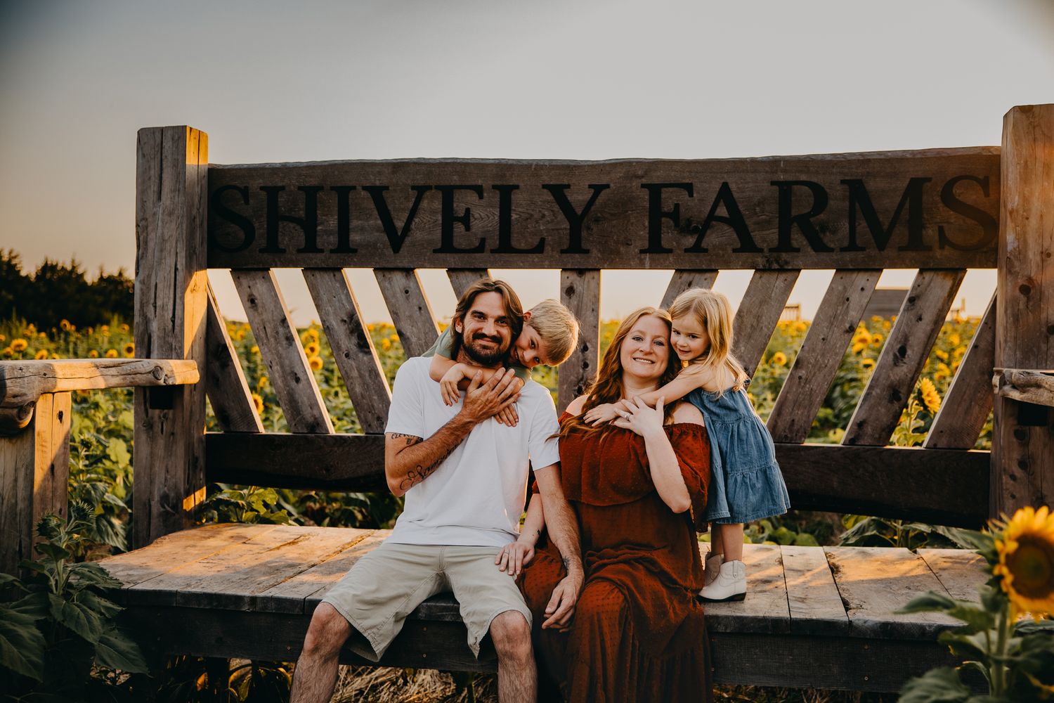 Family of three smiling at Shively Farms sign surrounded by sunflowers at golden hour.