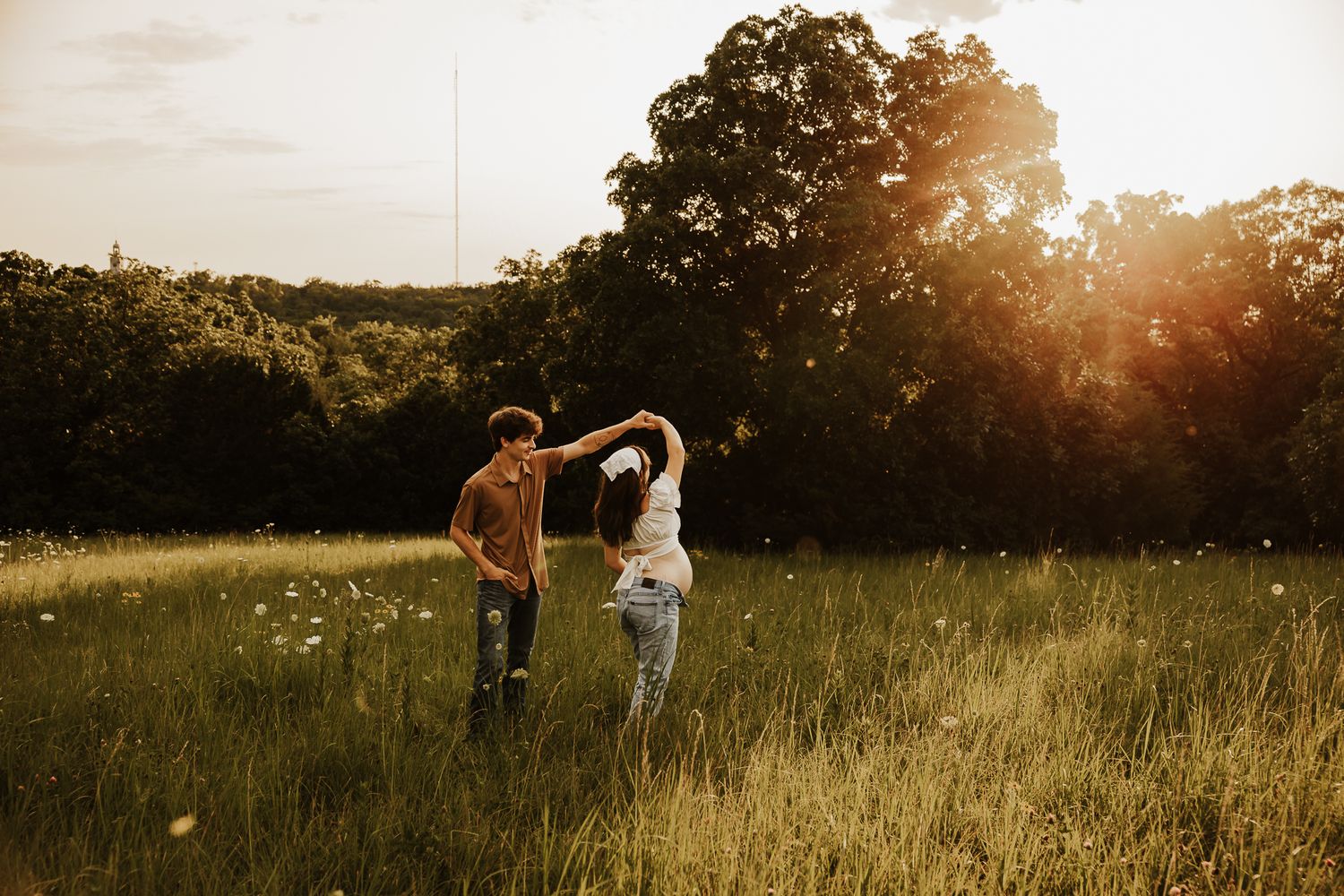 Couple dancing in a sunlit meadow surrounded by wildflowers and trees at golden hour.
