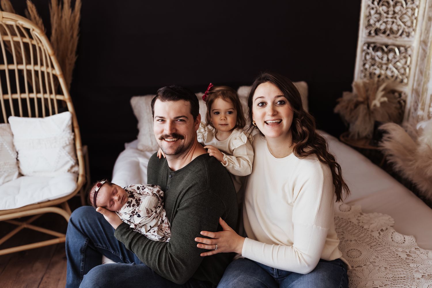 A happy family of four poses together, with dad holding a newborn baby and mom embracing a toddler girl in a cozy studio.