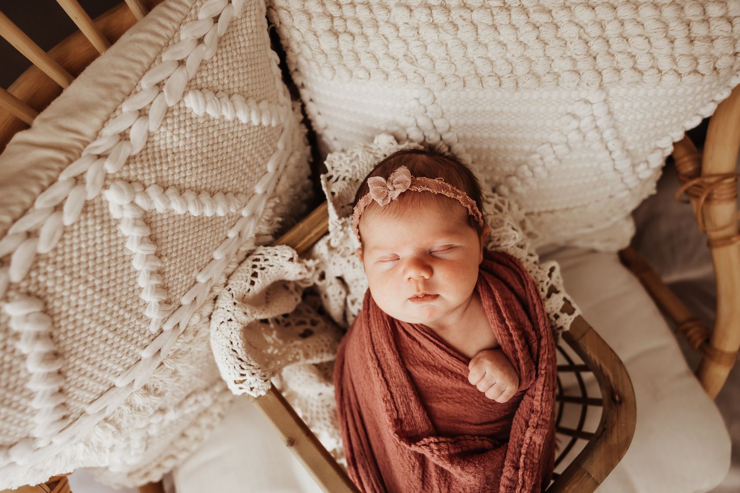 Newborn baby swaddled in rust wrap with floral headband, sleeping peacefully in a boho wicker basket.
