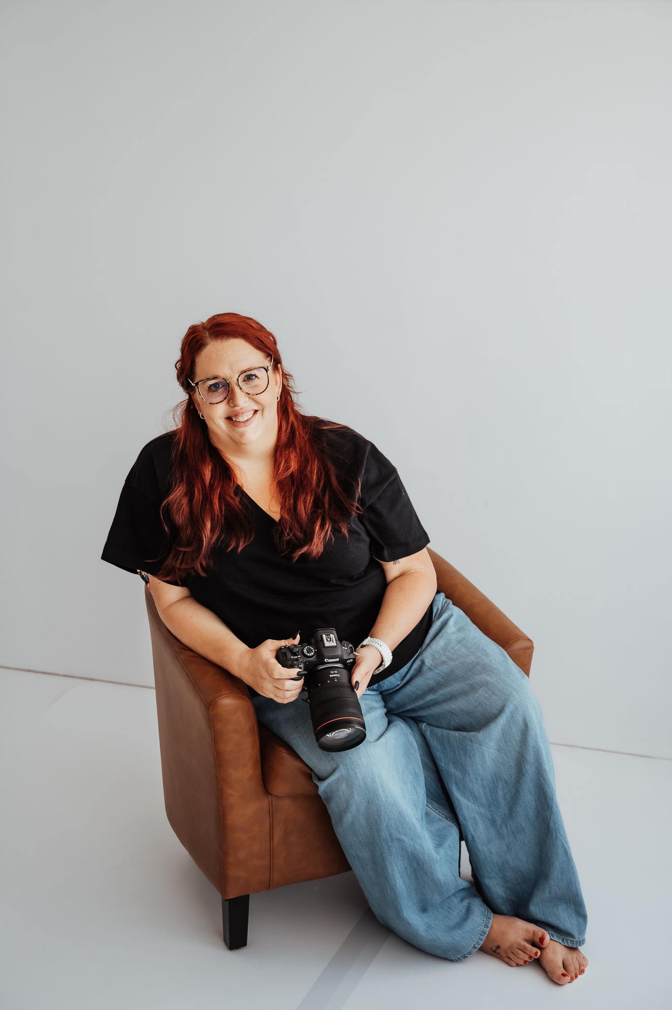 Red-haired female photographer sitting in leather chair holding DSLR camera, wearing black shirt and jeans.
