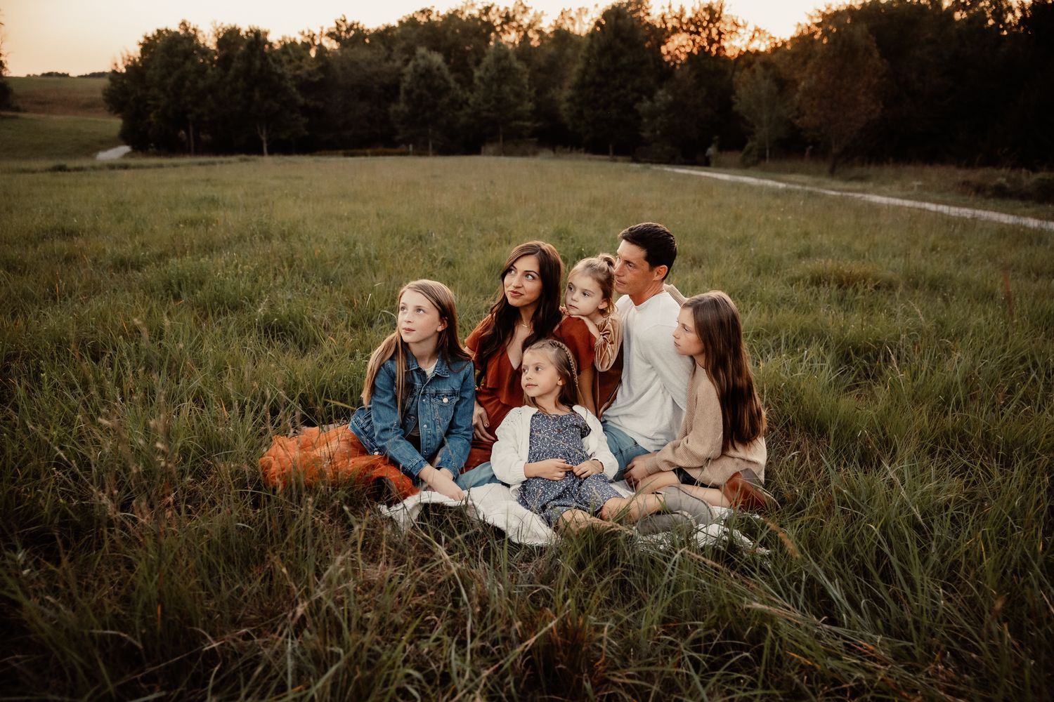 Family of five sitting together in a lush green field at golden hour sunset, smiling and enjoying a peaceful outdoor moment.