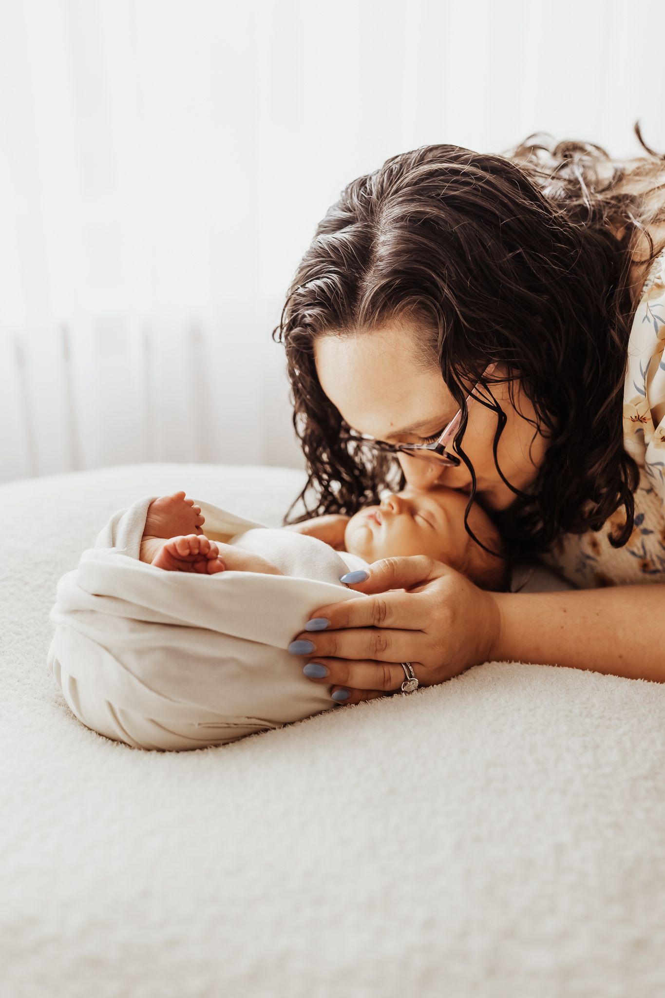 Loving mother gently kissing her sleeping newborn baby wrapped in a white swaddle on a soft white bed.
