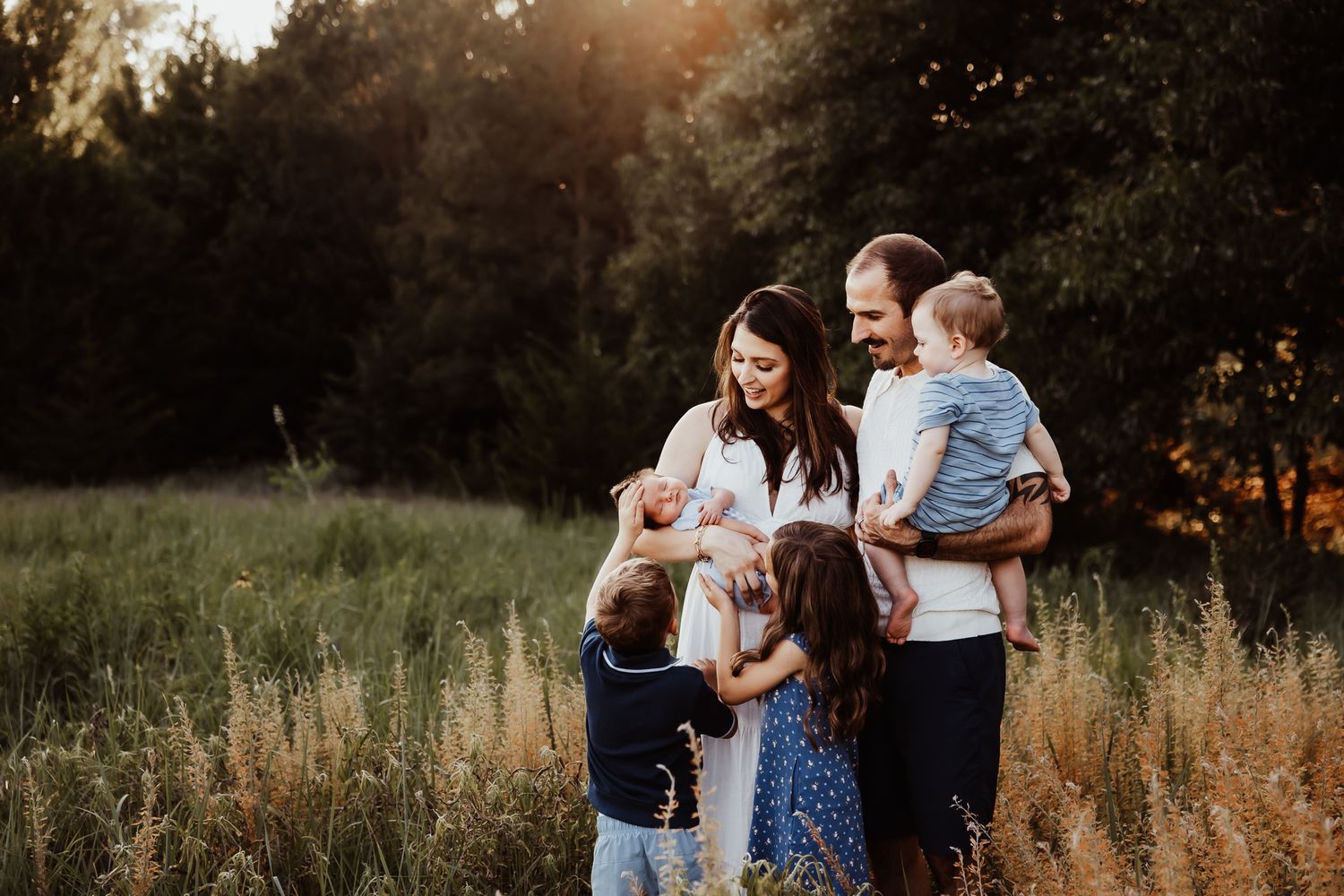Family of five sharing a warm moment in a sunlit meadow surrounded by lush green trees at golden hour.