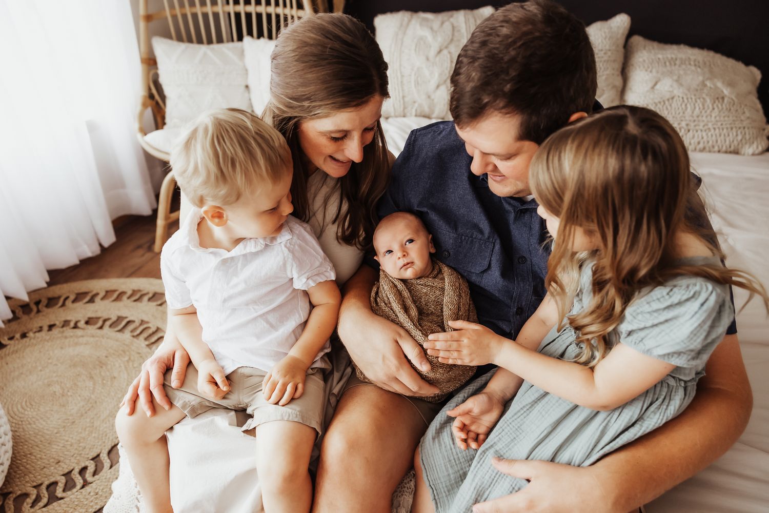 Happy family of five sitting on a bed, parents smiling at newborn baby held by father, with two young children.