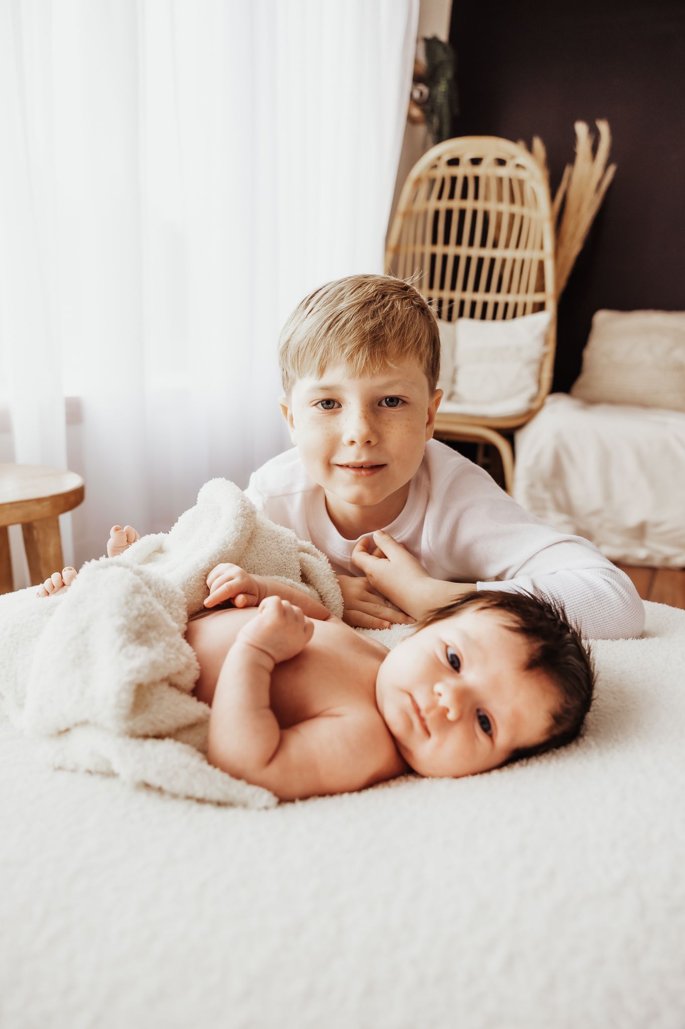 Young boy in white shirt lying on bed with newborn baby sibling wrapped in soft white blanket indoors.