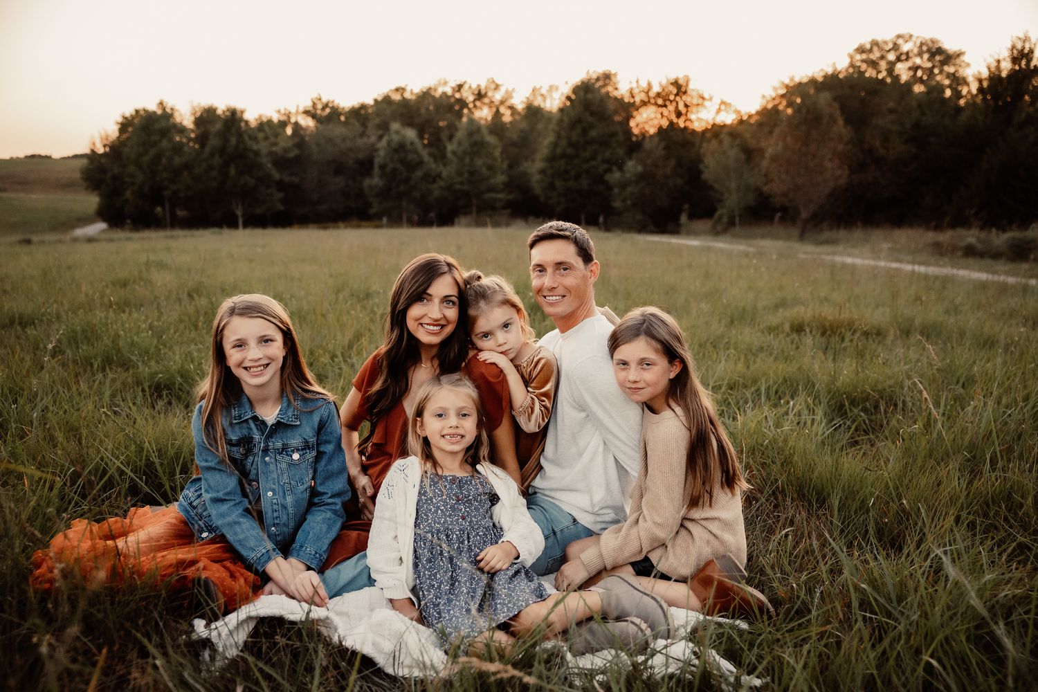 A happy family of six poses together in a lush green field at golden hour, smiling warmly for a portrait.