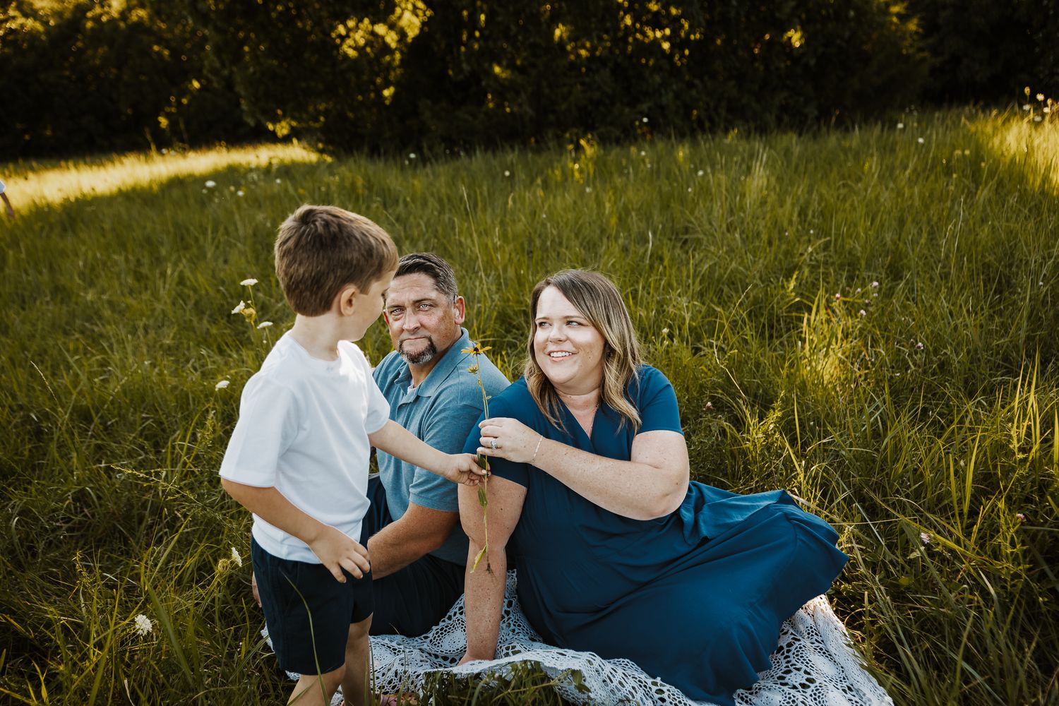 Family of three enjoying a sunny outdoor picnic in a lush green meadow, mother in teal dress smiling at young son.