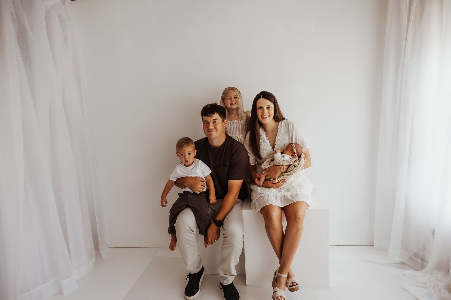 A happy family of four poses in a bright white studio, with parents holding two young children including a newborn.