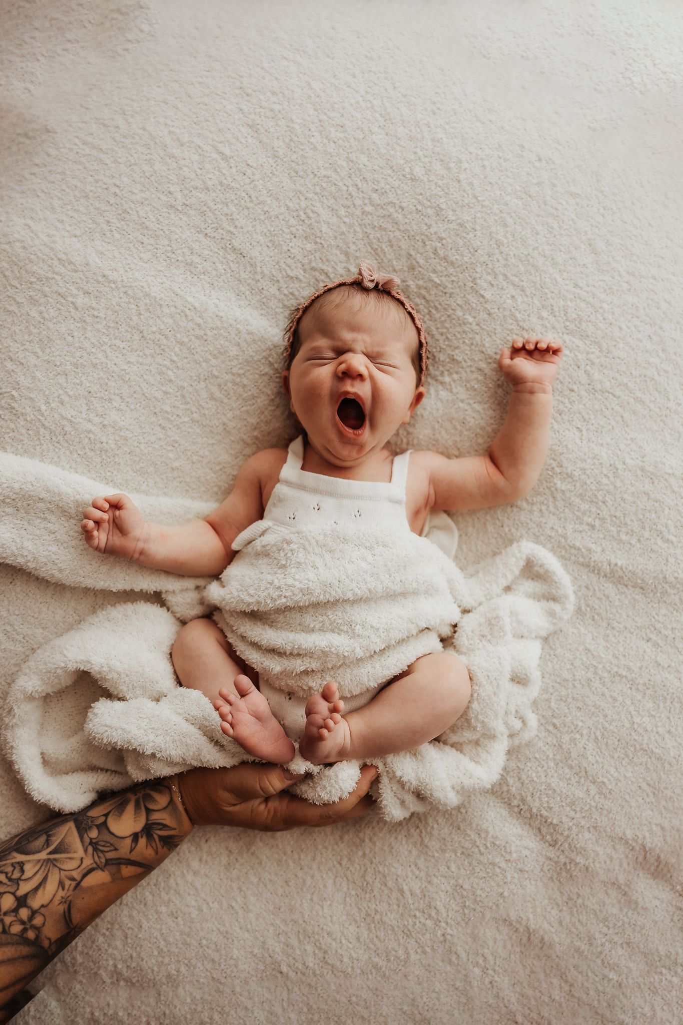 Newborn baby yawning and stretching on a white blanket, wearing a white romper with a pink headband.