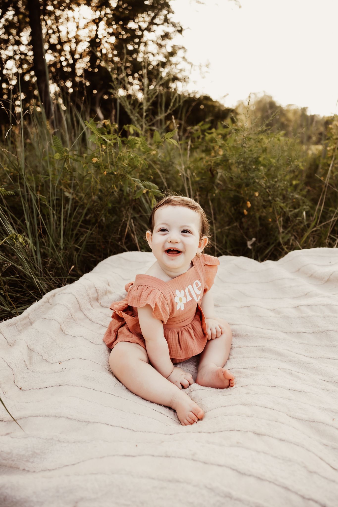 Smiling baby girl in rust romper sitting on white blanket outdoors in golden hour light.