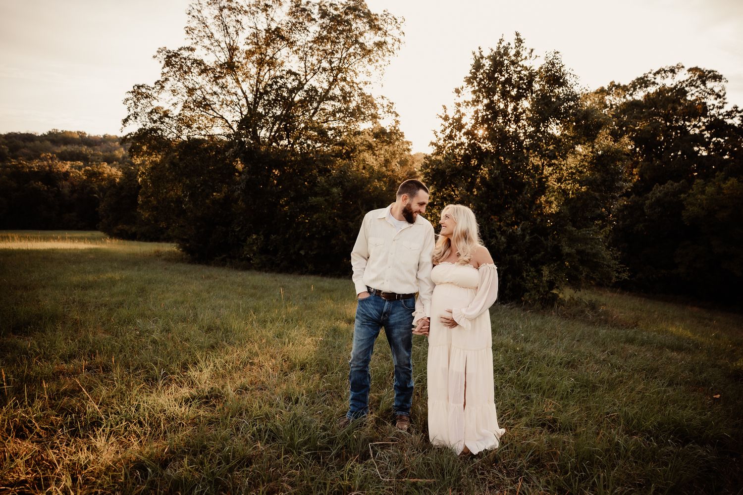 Couple sharing a tender moment in a sunlit field at golden hour, woman in white maternity gown, man in jeans and white sweater.