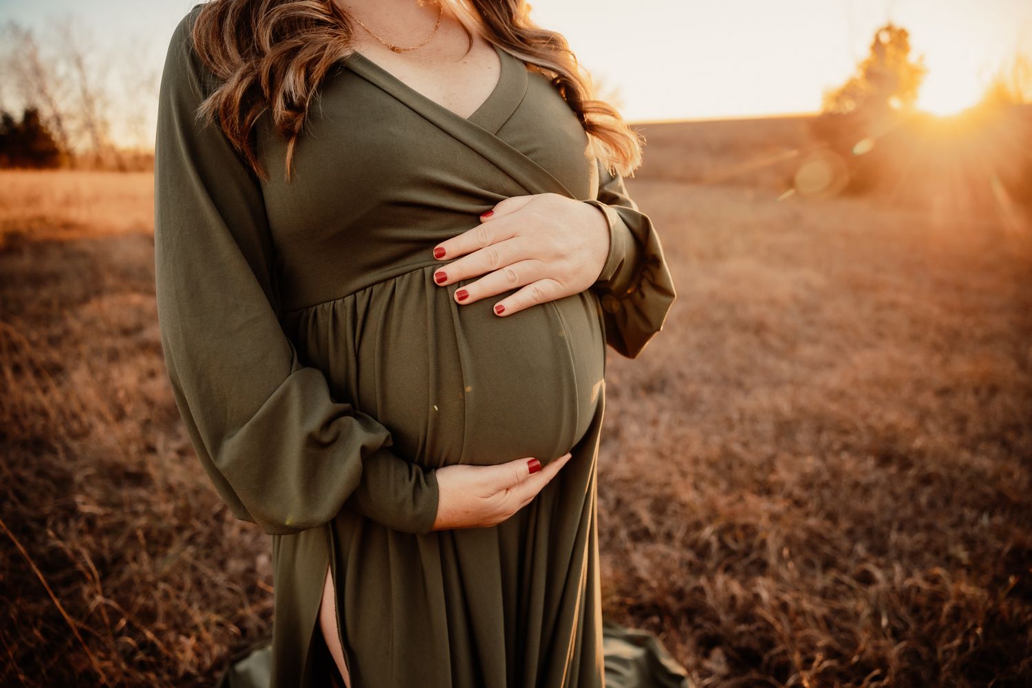 Pregnant woman in olive green dress cradling baby bump at golden hour in a field.