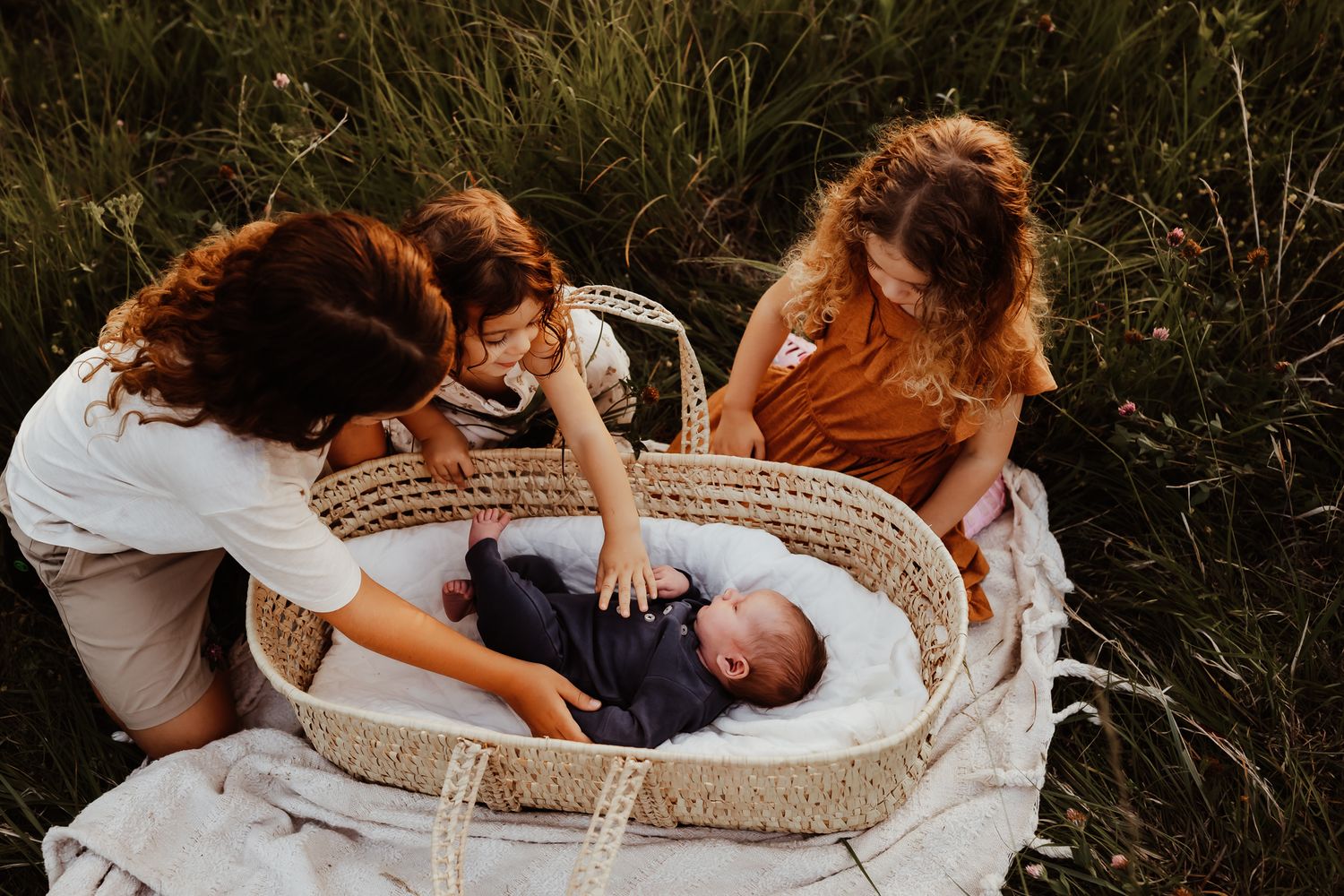 Mother and two young girls admiring a newborn baby in a wicker Moses basket outdoors in a green meadow.