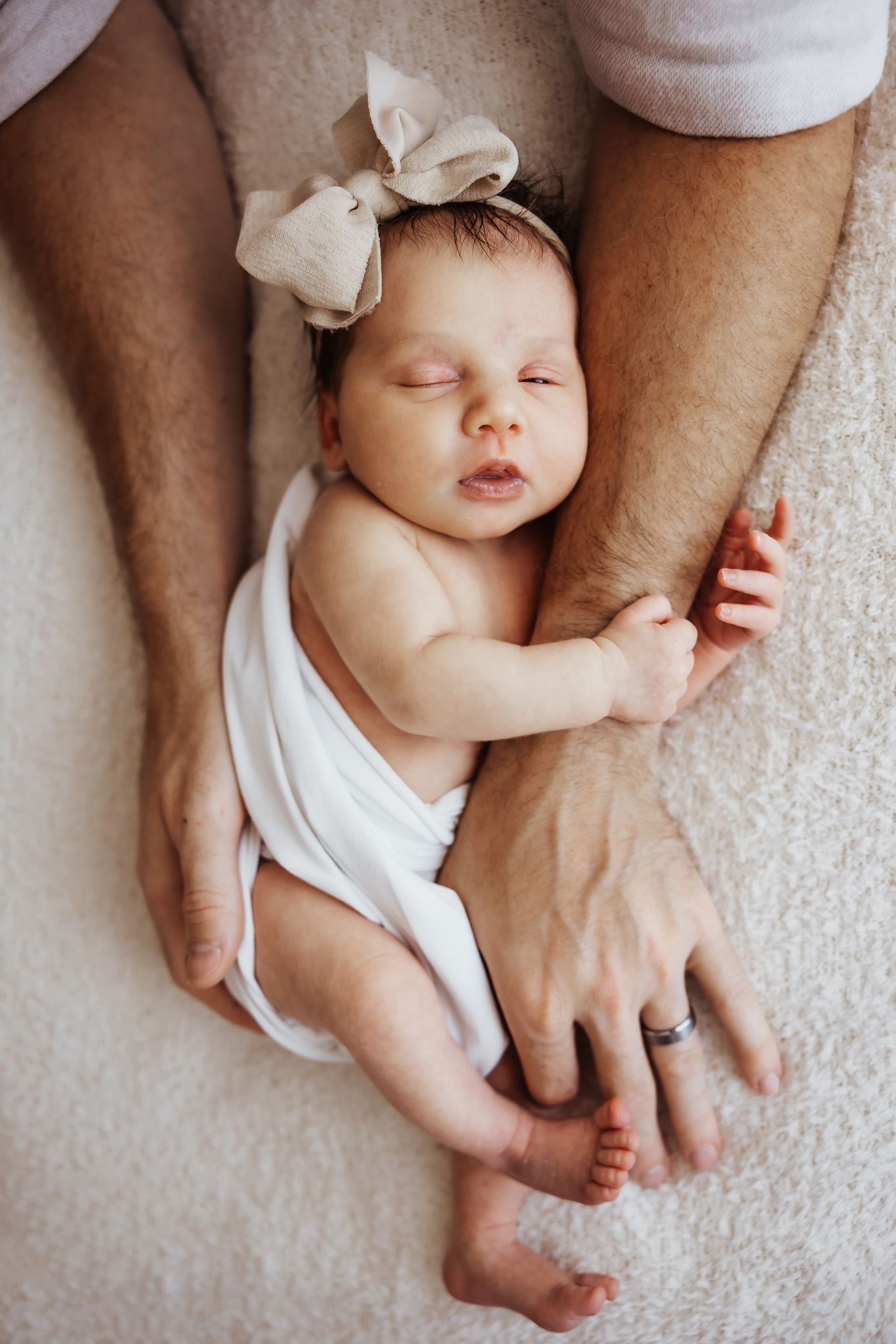 Sleeping newborn baby girl with bow headband rests peacefully between her father's arms on a soft blanket.