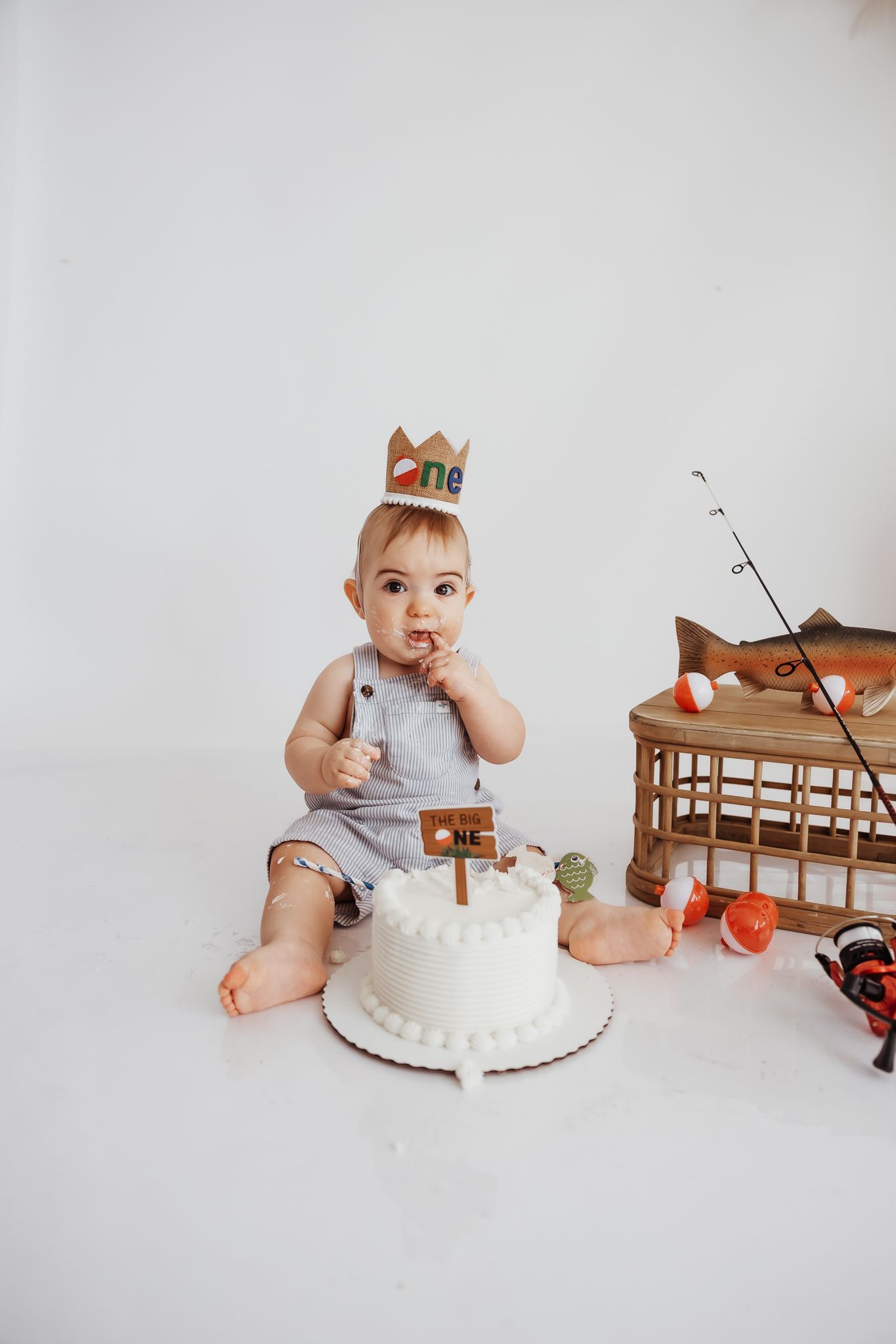 Baby boy in gray romper wearing a crown celebrates first birthday with a white smash cake in a white studio.
