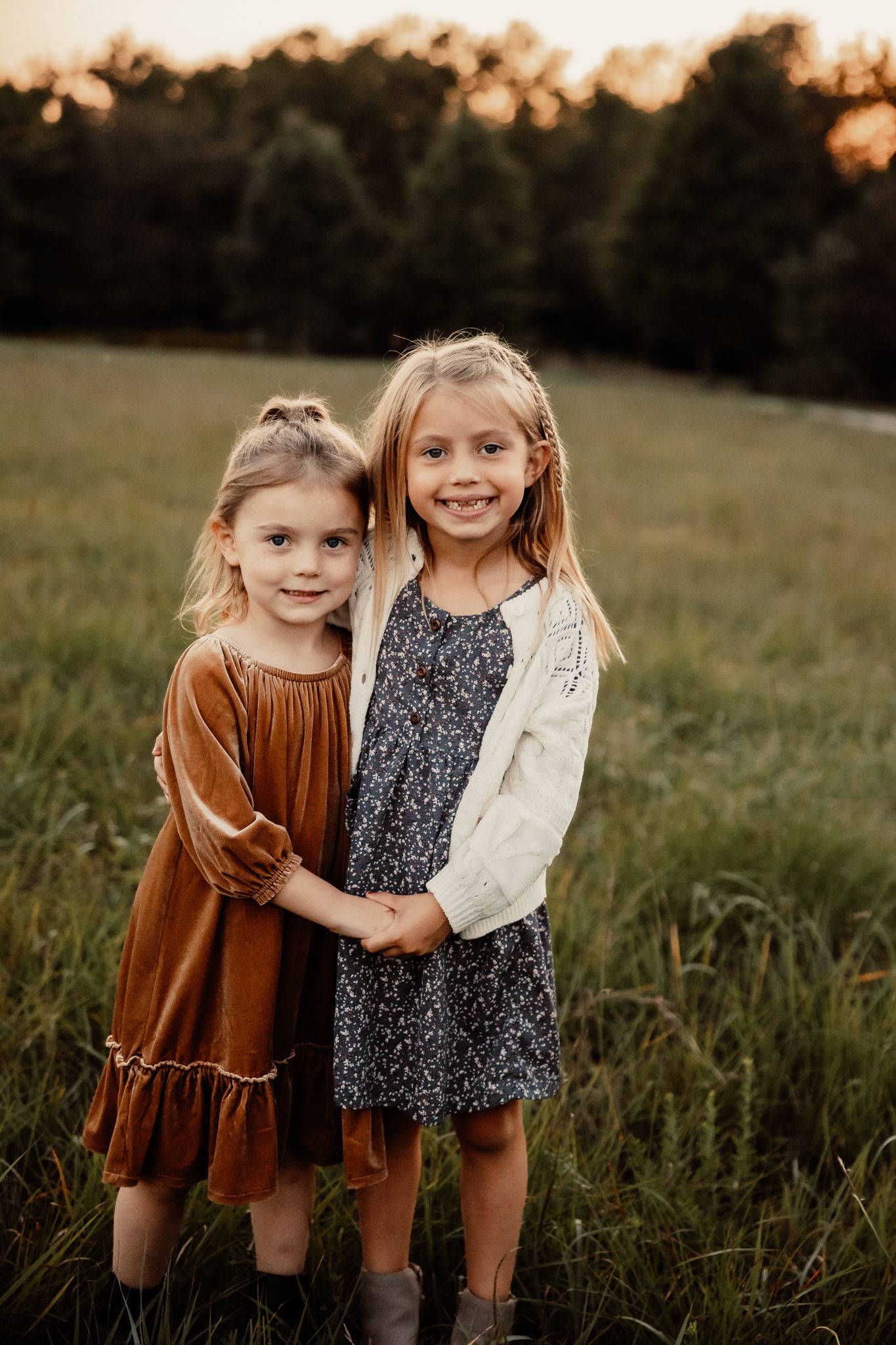 Two young girls in autumn dresses embrace in a grassy field at golden hour sunset.