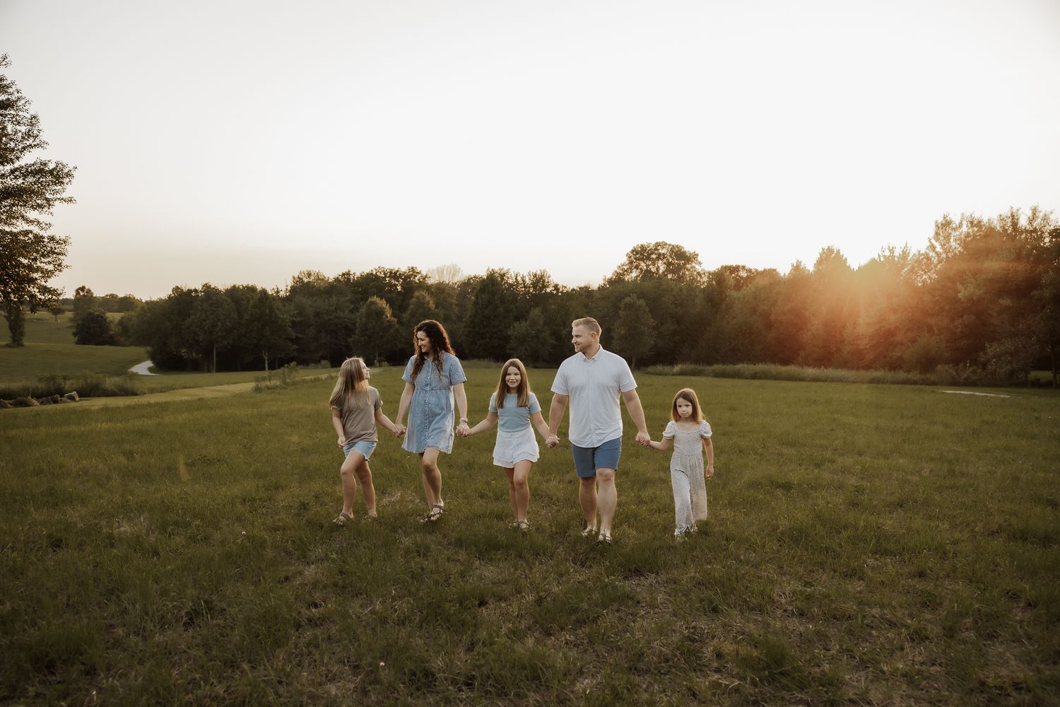 Family of five walking hand-in-hand through a green field during golden hour sunset.