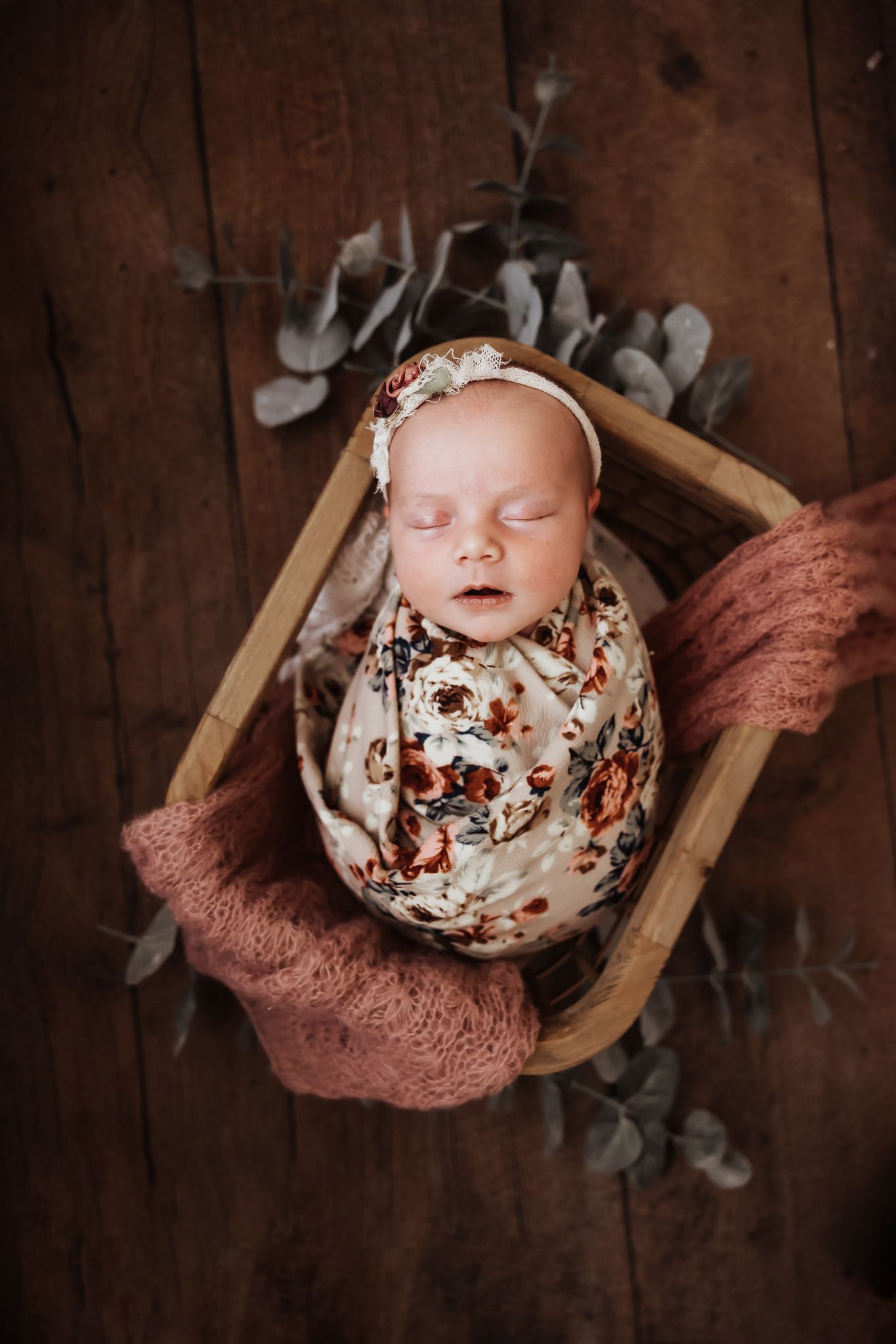 Sleeping newborn baby swaddled in floral wrap with pink knit blanket in wooden basket on rustic floor.