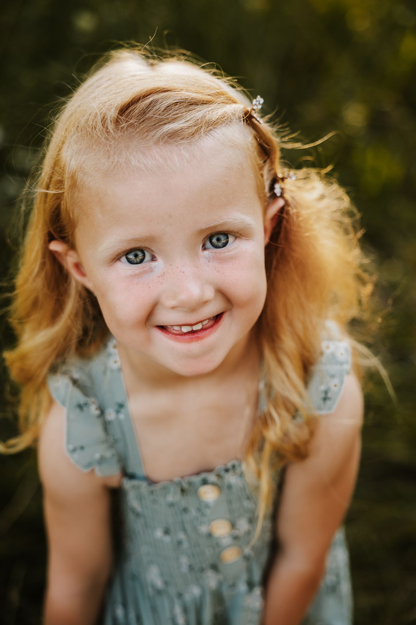 A smiling young girl with red hair and green eyes wearing a sage green dress outdoors.