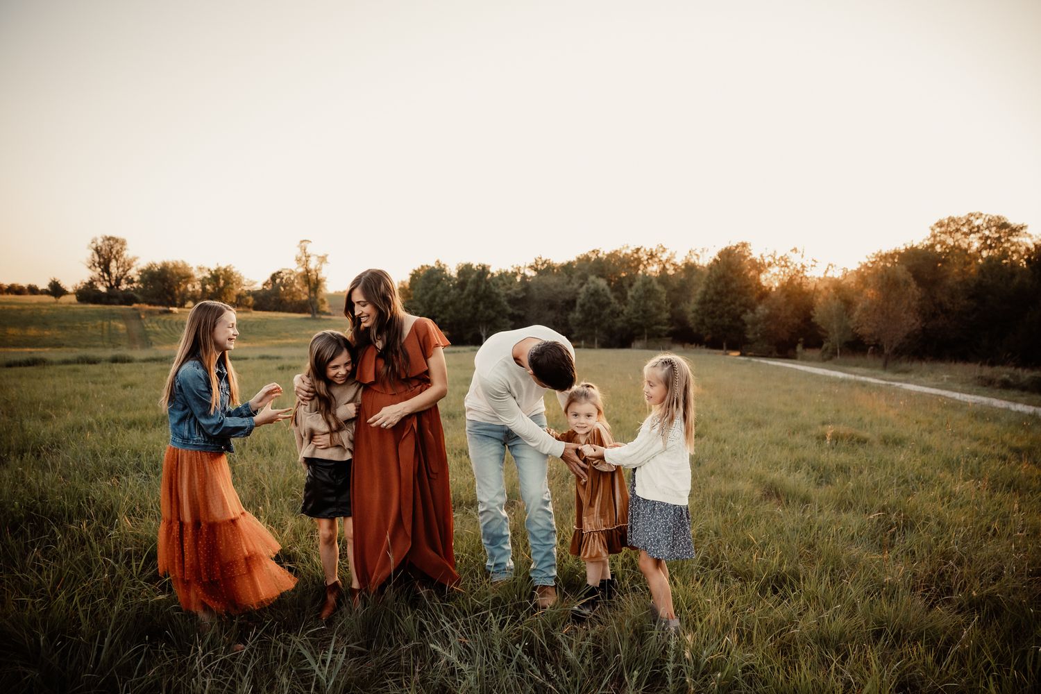 Family of five enjoying a golden hour sunset in a green meadow, wearing warm earth tones, sharing a joyful moment together.