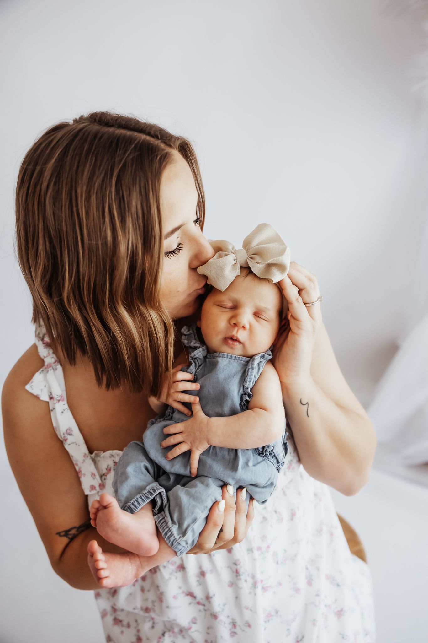 A mother tenderly kisses her sleeping newborn baby girl wearing a bow headband and blue romper.