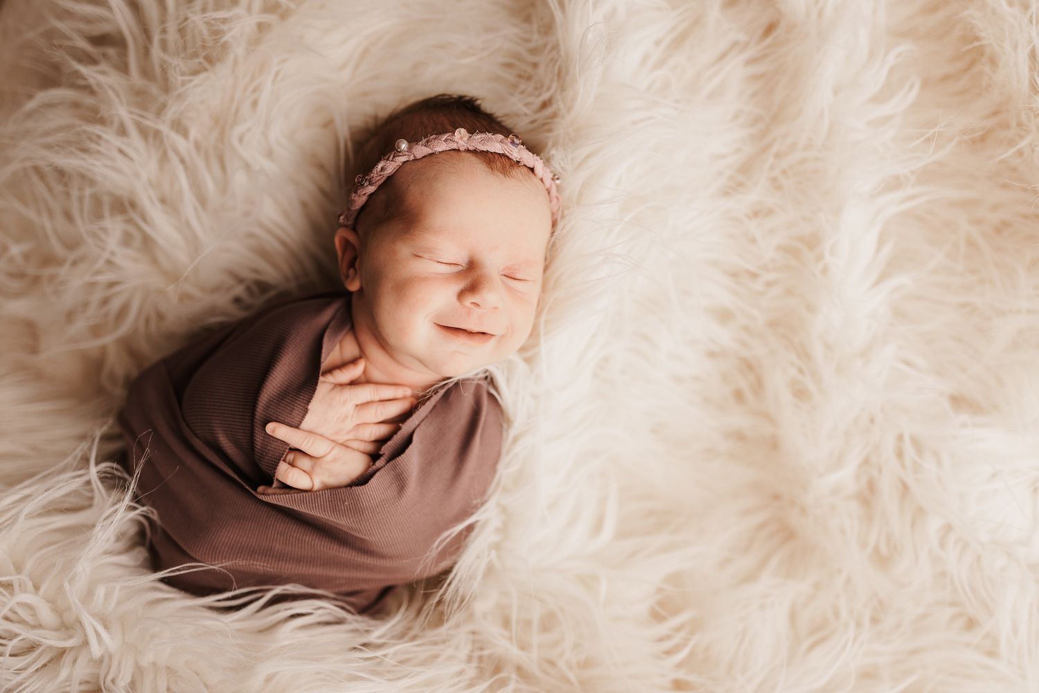 Sleeping newborn baby swaddled in brown wrap with pink floral headband on fluffy white fur blanket.