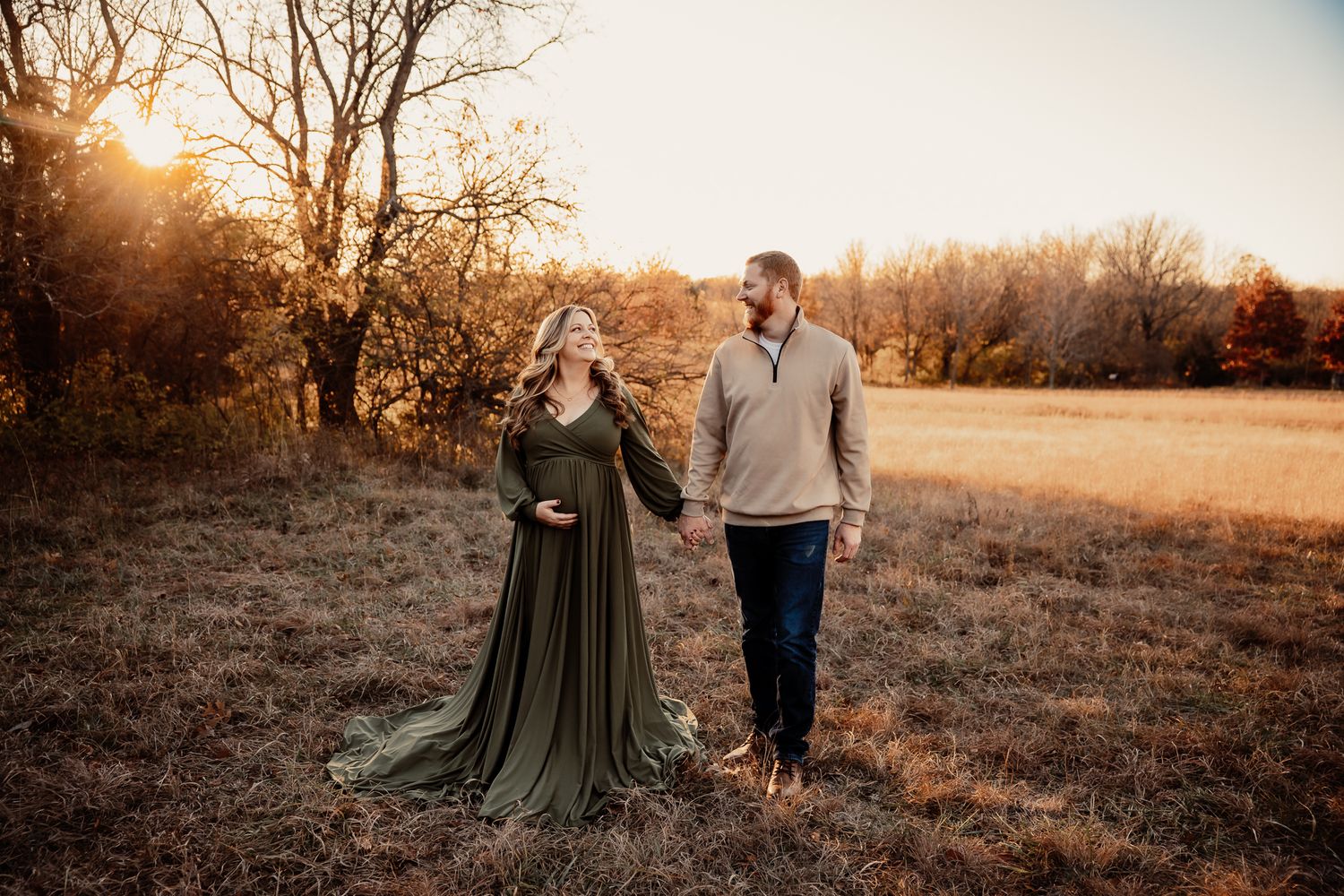 Couple holding hands in golden sunset light, woman in flowing olive maternity gown, man in casual attire, outdoor fall field.