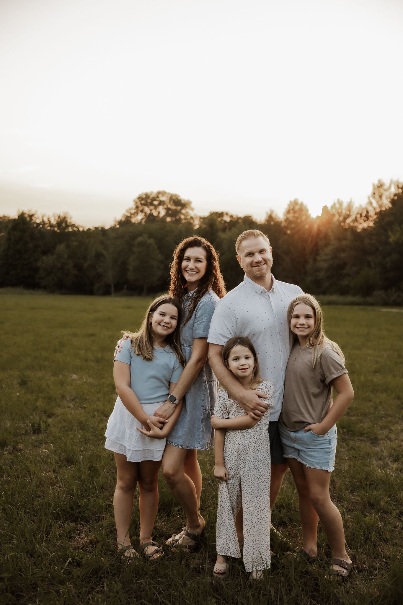 Family of five posing in a sunlit field at golden hour, parents with three daughters smiling outdoors.