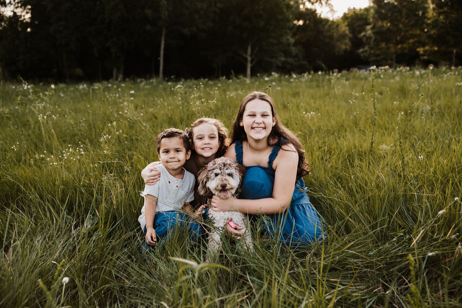 Three girls and a dog sitting in a lush green meadow, smiling during golden hour outdoor family photo.