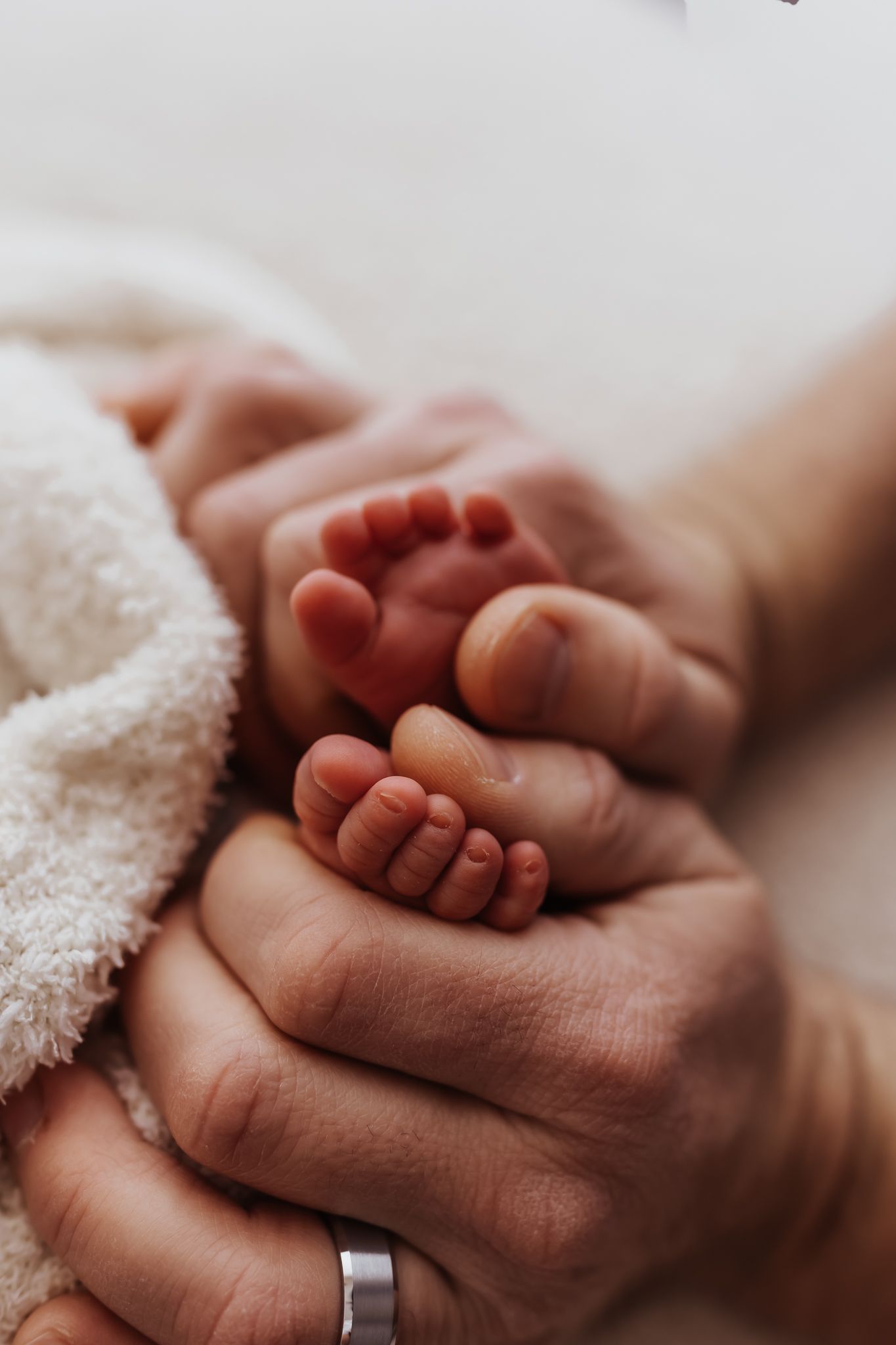 Adult hands gently cradling tiny newborn baby feet wrapped in a soft white blanket, with a wedding ring visible.