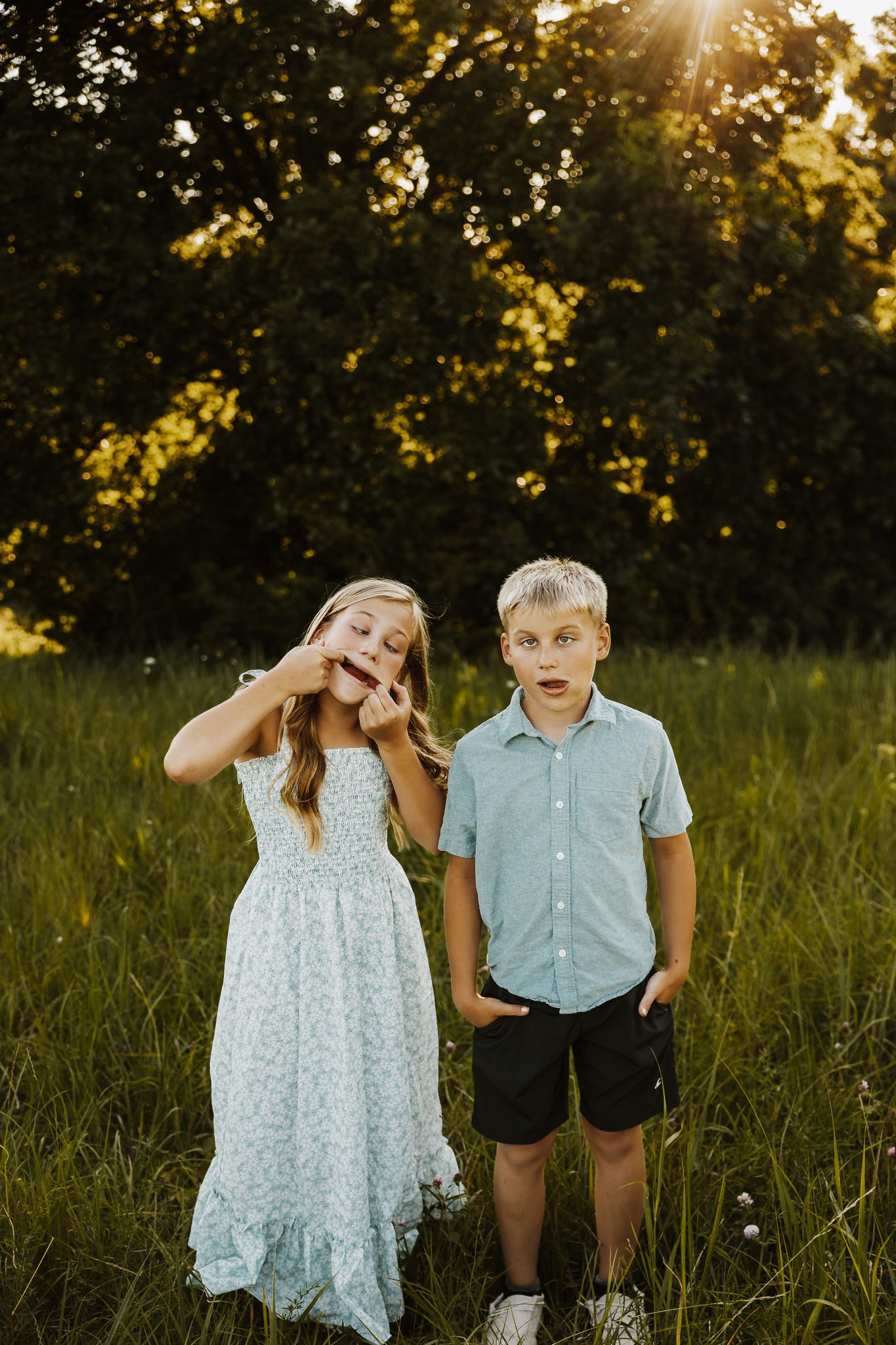 Two children pose in a sunlit field at golden hour, girl in floral dress making faces, boy in blue shirt looking surprised.