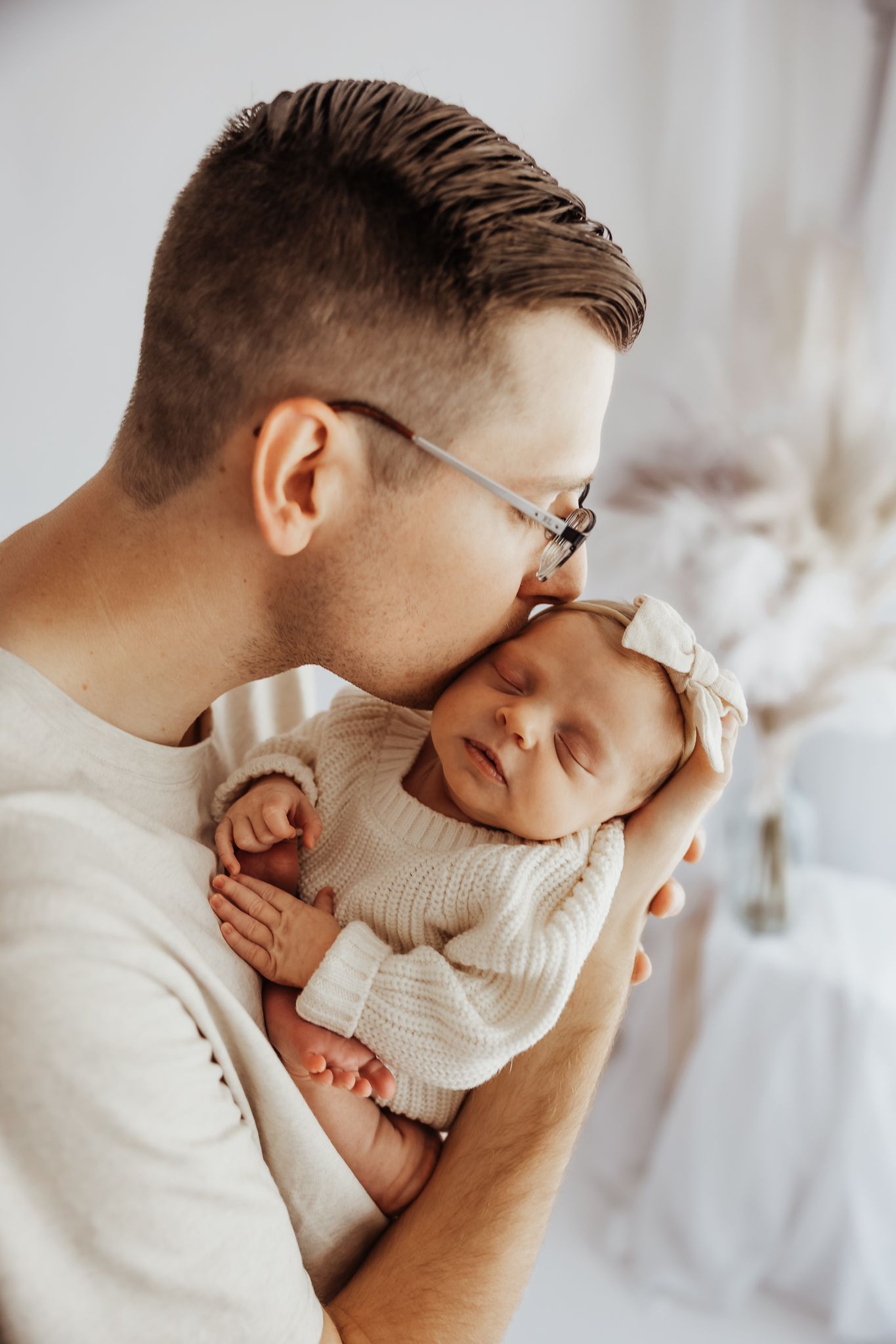 Father in glasses tenderly kissing sleeping newborn baby girl in white knit outfit.