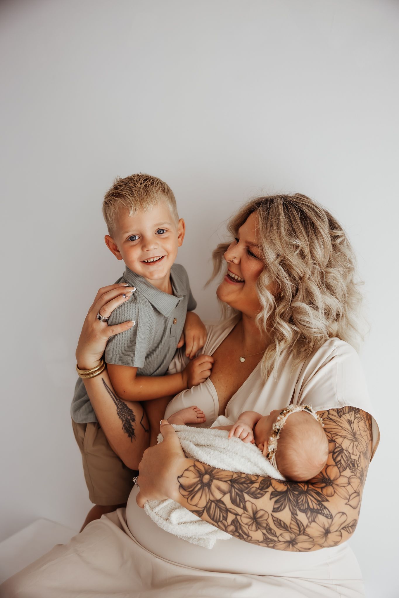 Smiling tattooed blonde mother holding newborn baby while young blond boy poses beside her.