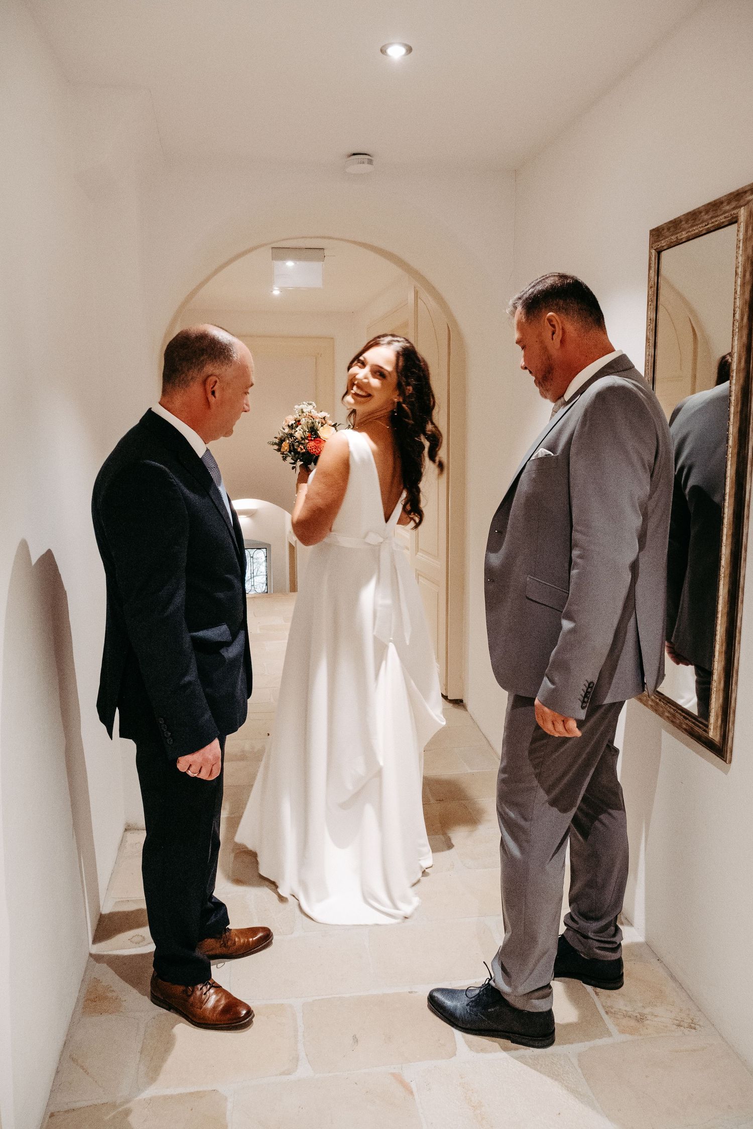 Smiling bride in white dress holding bouquet chatting with two male guests indoors.