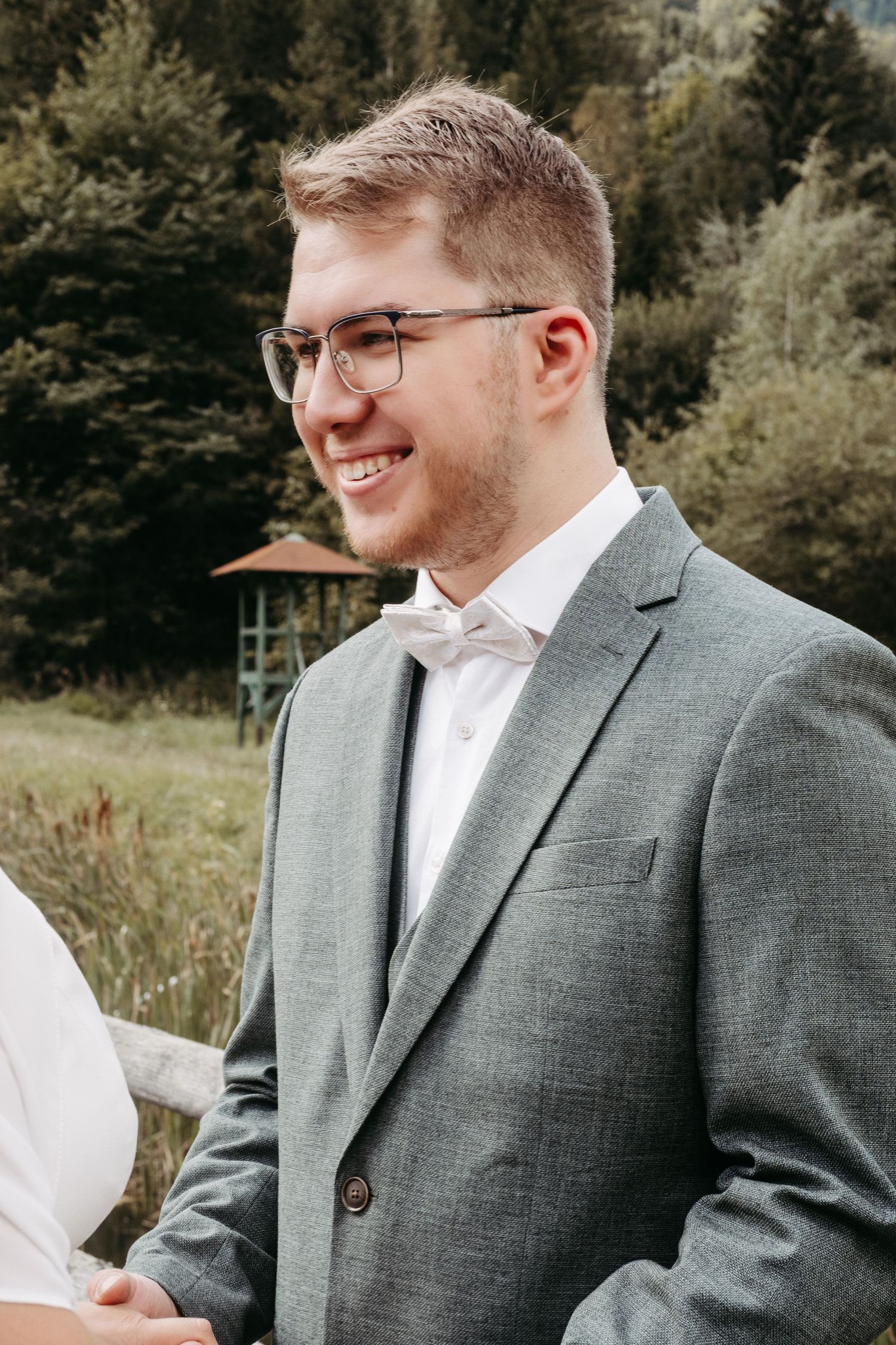 Smiling groom in gray suit and bow tie standing outdoors at a wedding ceremony with forest background.