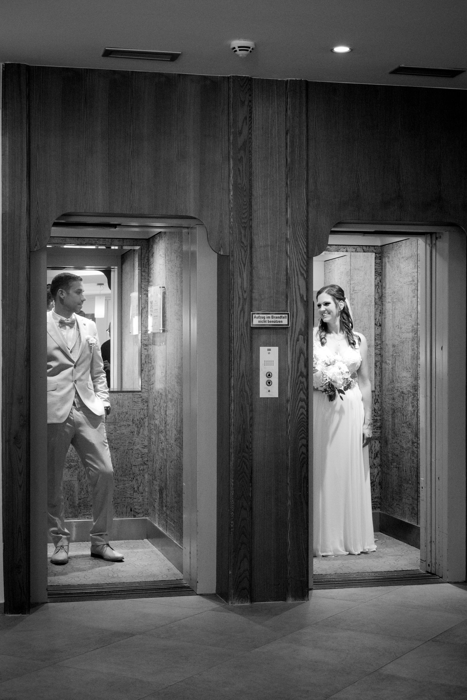 Black and white photo of bride and groom standing in separate elevator doors at their wedding venue.