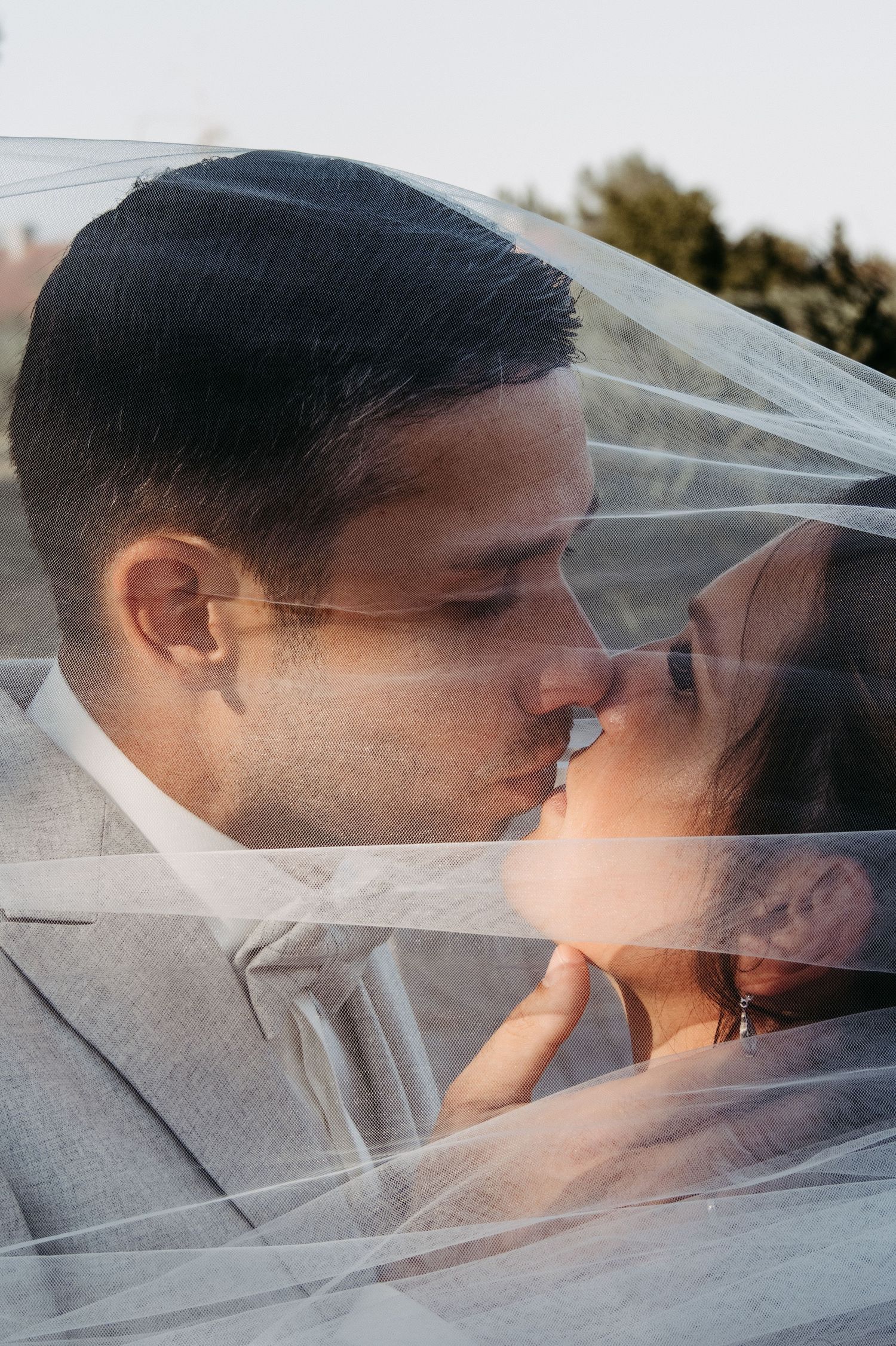 A bride and groom share a romantic kiss under a flowing white wedding veil in a soft golden light.