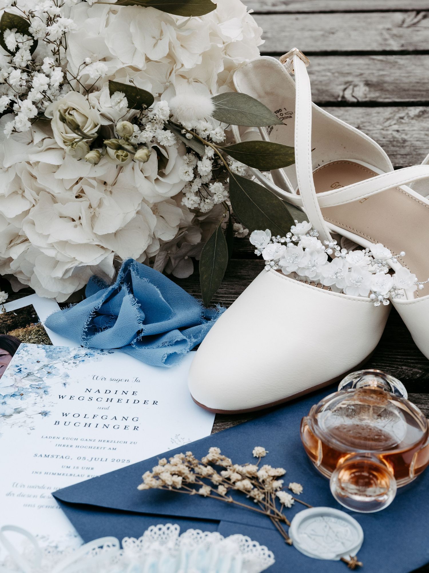 Elegant wedding flat lay with white bridal shoes, hydrangea bouquet, blue invitation, perfume bottle, and dried flowers on dark wood.