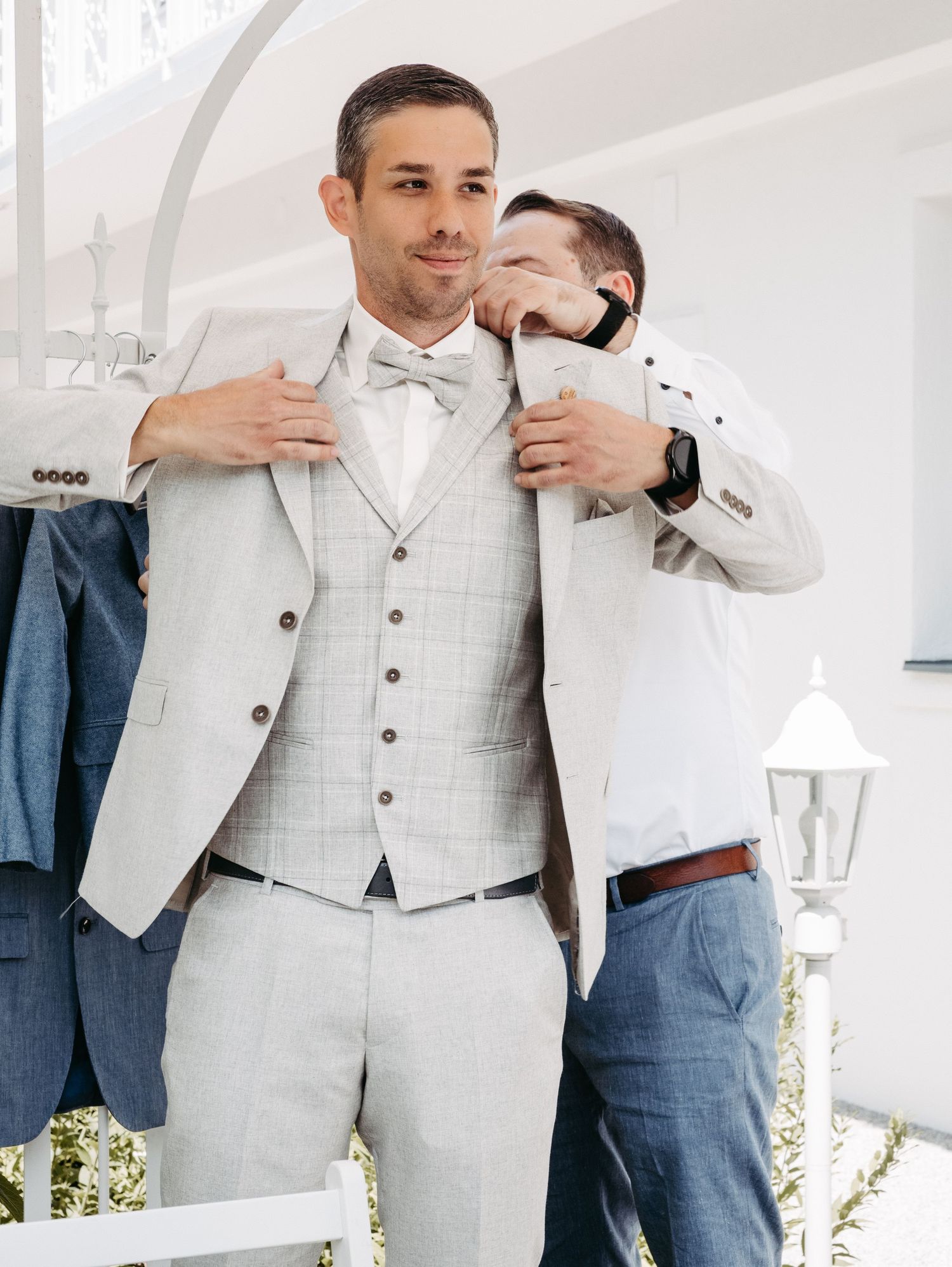 Groom in light gray three-piece suit being helped by groomsman adjusting jacket before wedding ceremony.