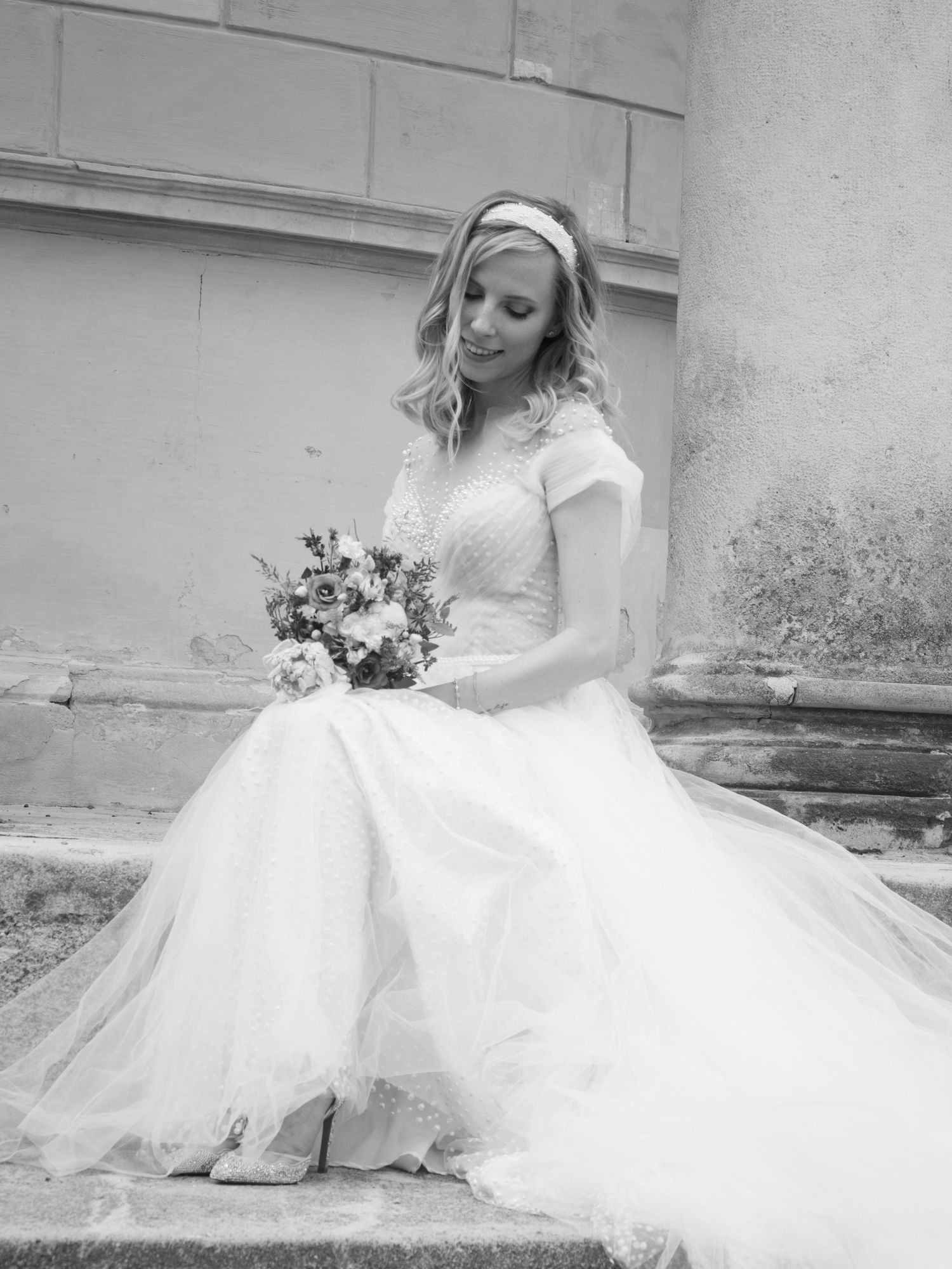 Bride in white wedding gown holding bouquet, seated outdoors in black and white portrait photo.
