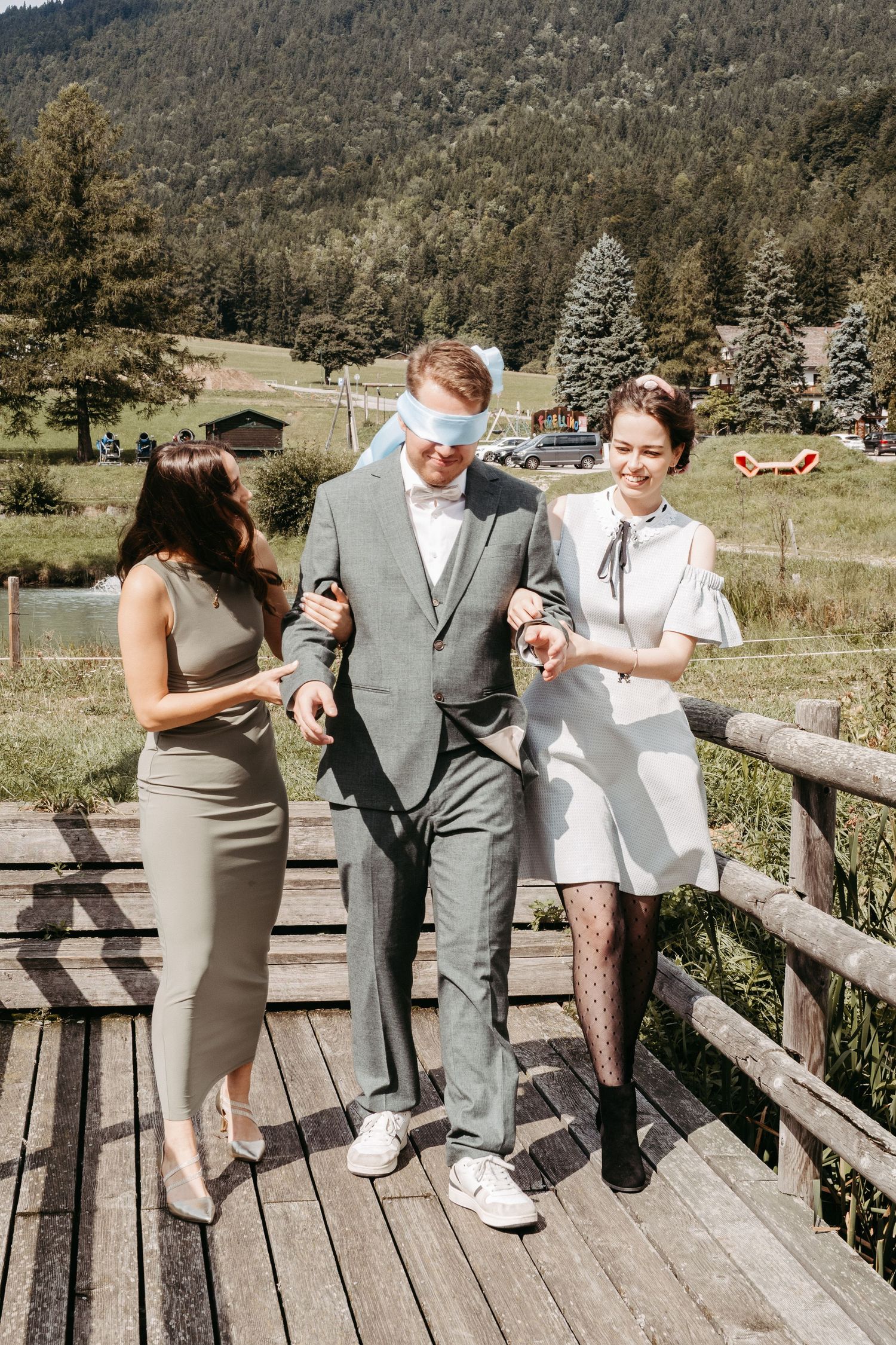 Blindfolded groom guided by two women on wooden bridge with mountain backdrop at outdoor wedding.