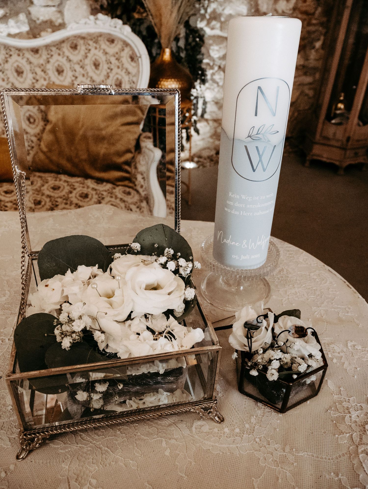 Elegant wedding unity candle with NW monogram beside white floral arrangements in glass boxes on lace tablecloth.