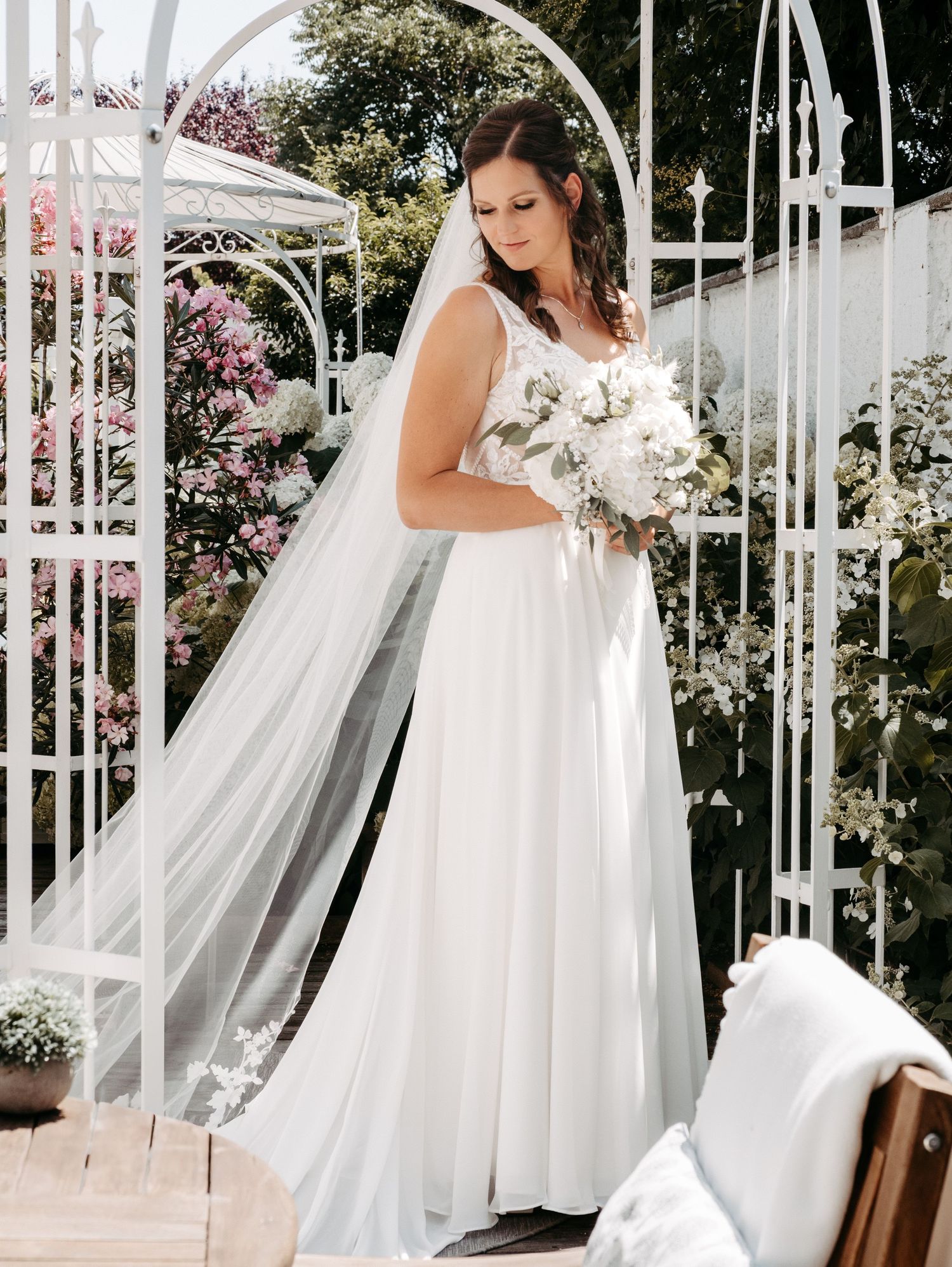 Bride in a flowing white wedding gown with veil holds a bouquet, standing in a white garden arch surrounded by blooms.