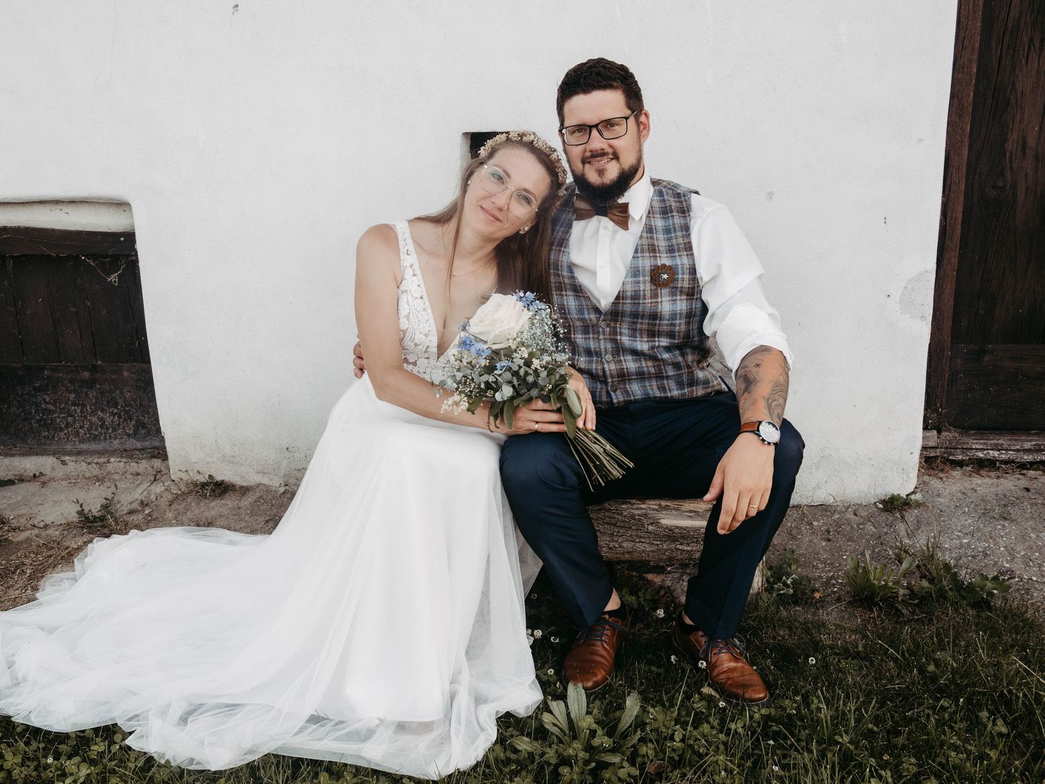 Bride in white gown holding bouquet leans on groom in plaid vest and bow tie seated on bench outdoors.