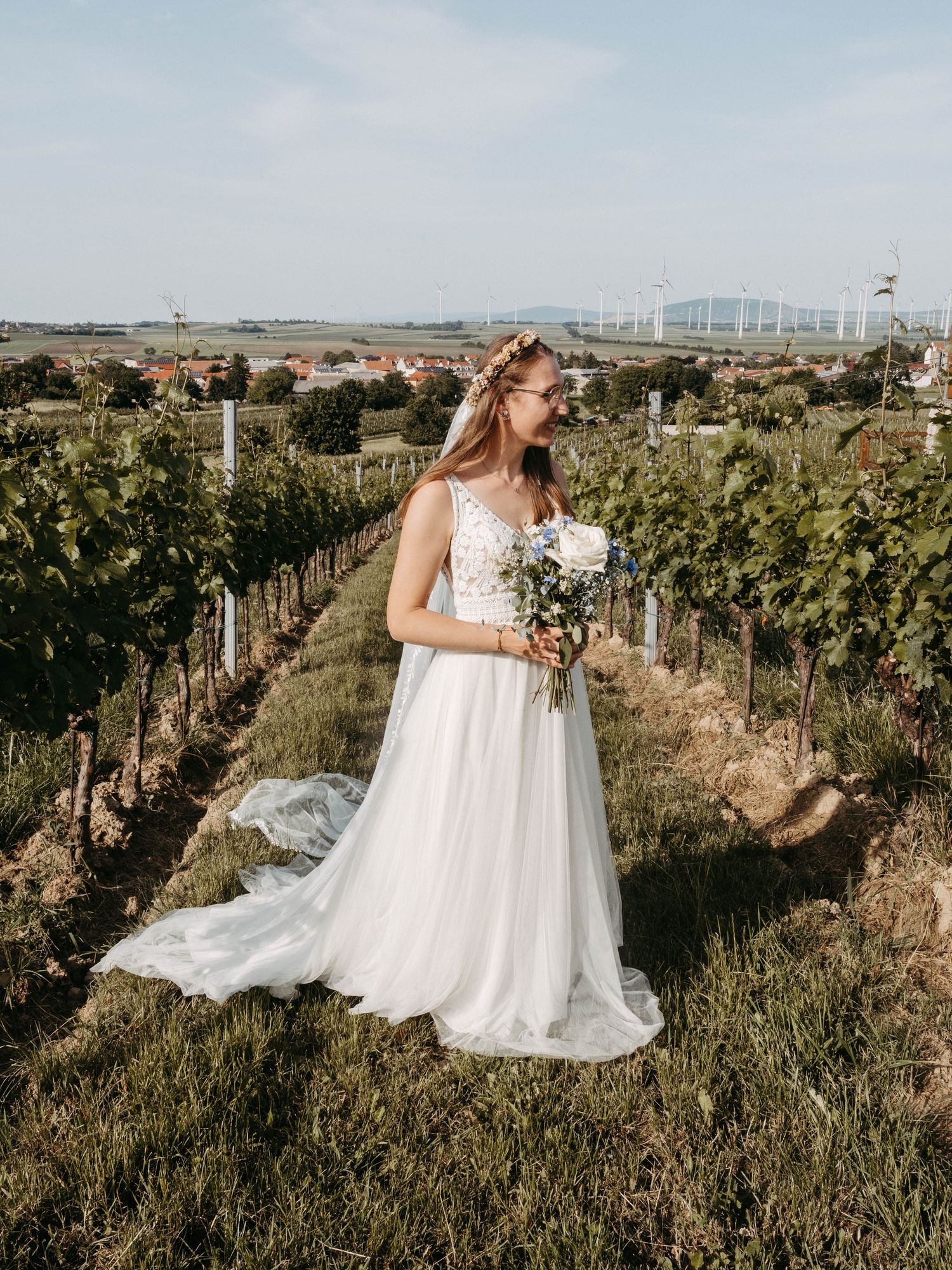Bride in white gown holding bouquet standing in vineyard rows with countryside backdrop on sunny day.