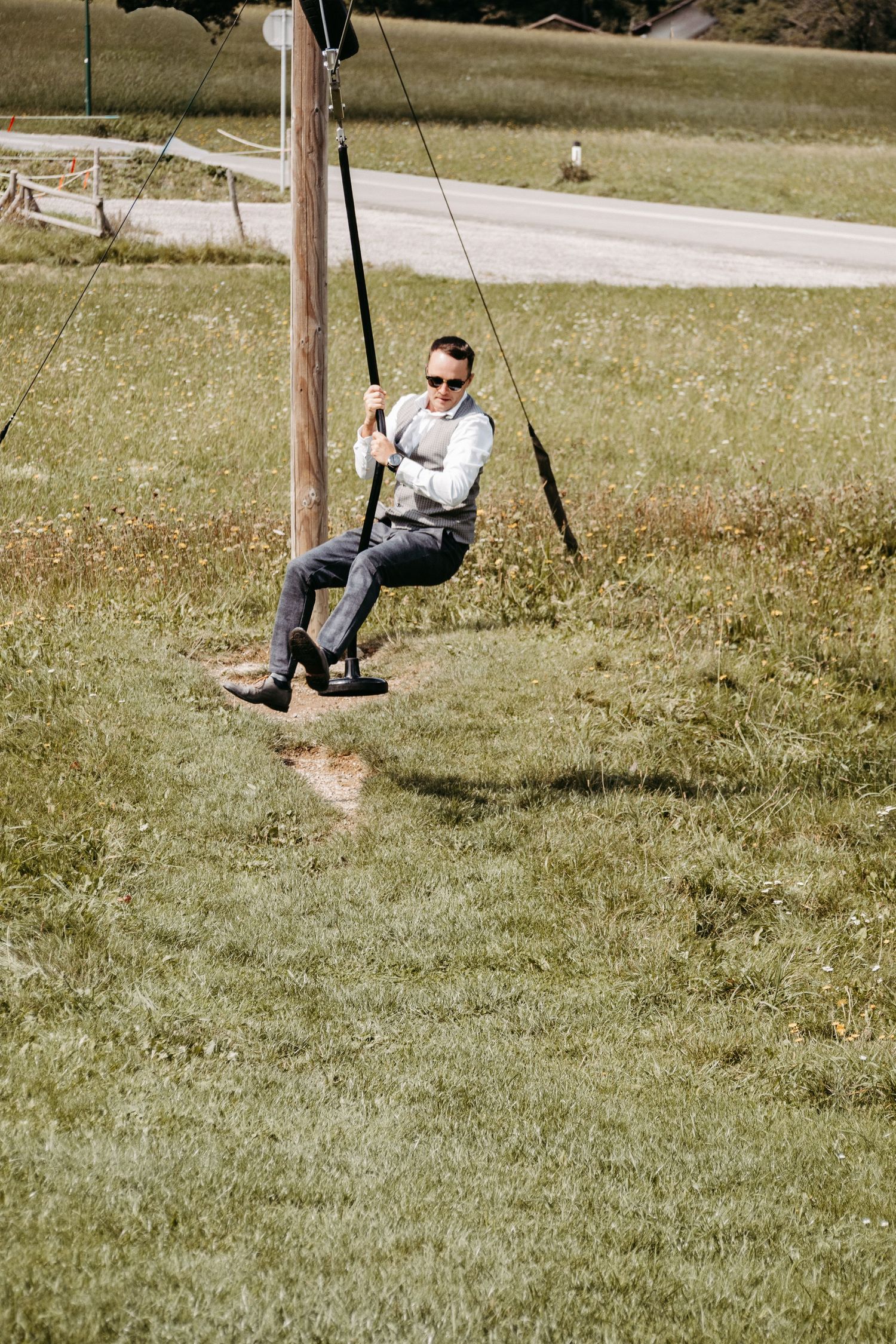 Man in sunglasses and dress clothes riding a zip line swing outdoors on a sunny day in a green field.