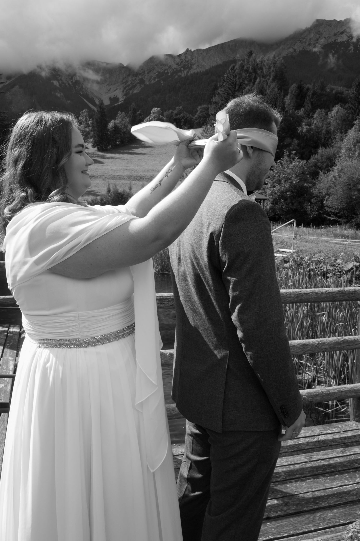 Bride tying a blindfold on groom outdoors with mountain scenery in black and white wedding photo.