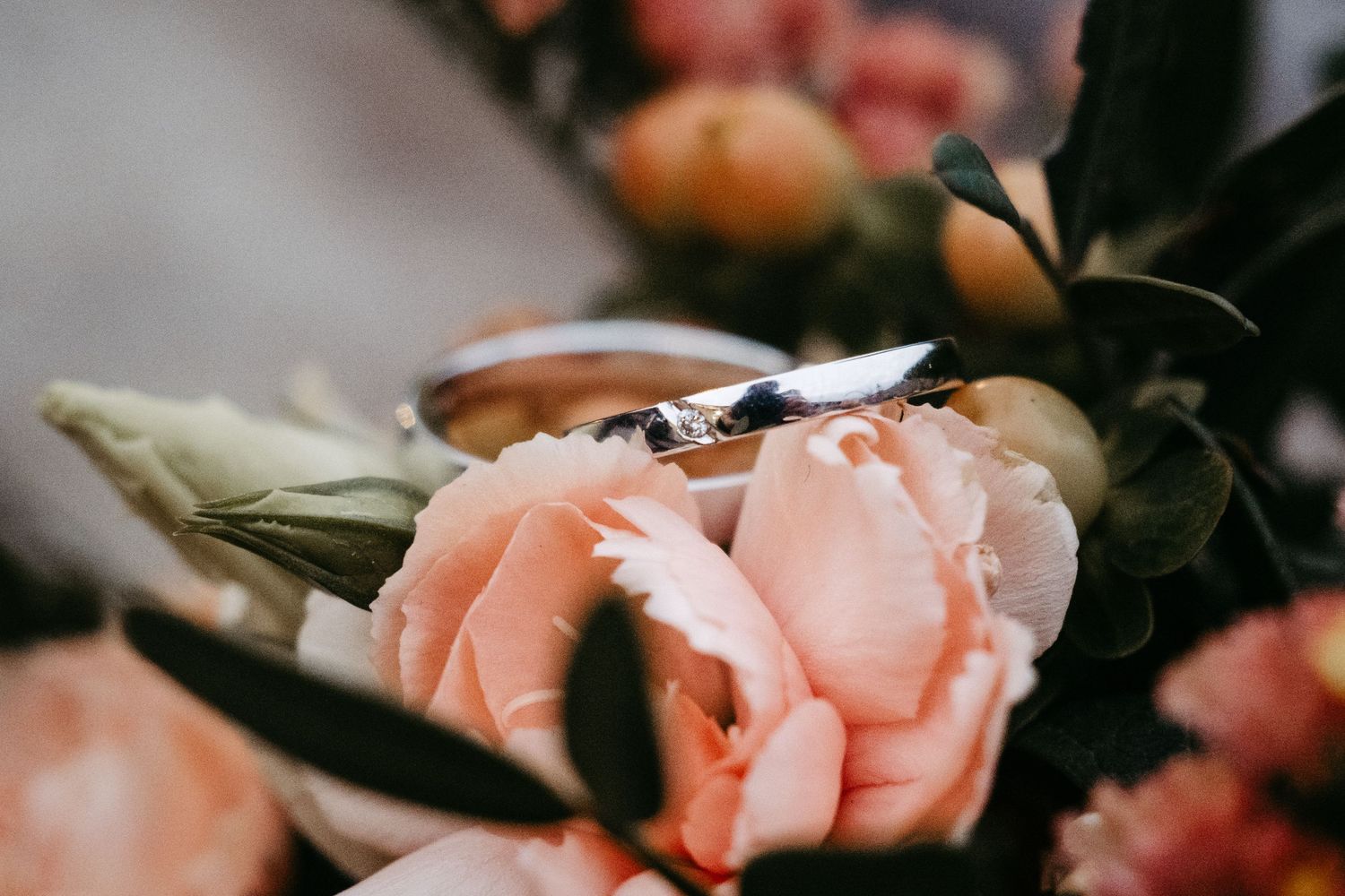 Two wedding rings resting on pink roses in a bridal bouquet with green leaves and orange accents.