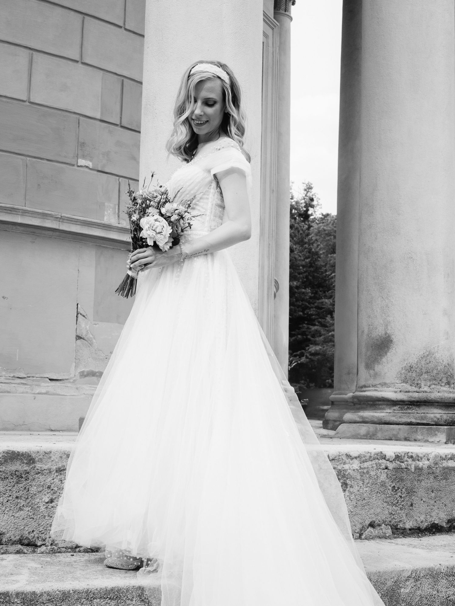 Black and white photo of a bride in a flowing tulle gown holding a bouquet beside stone columns.